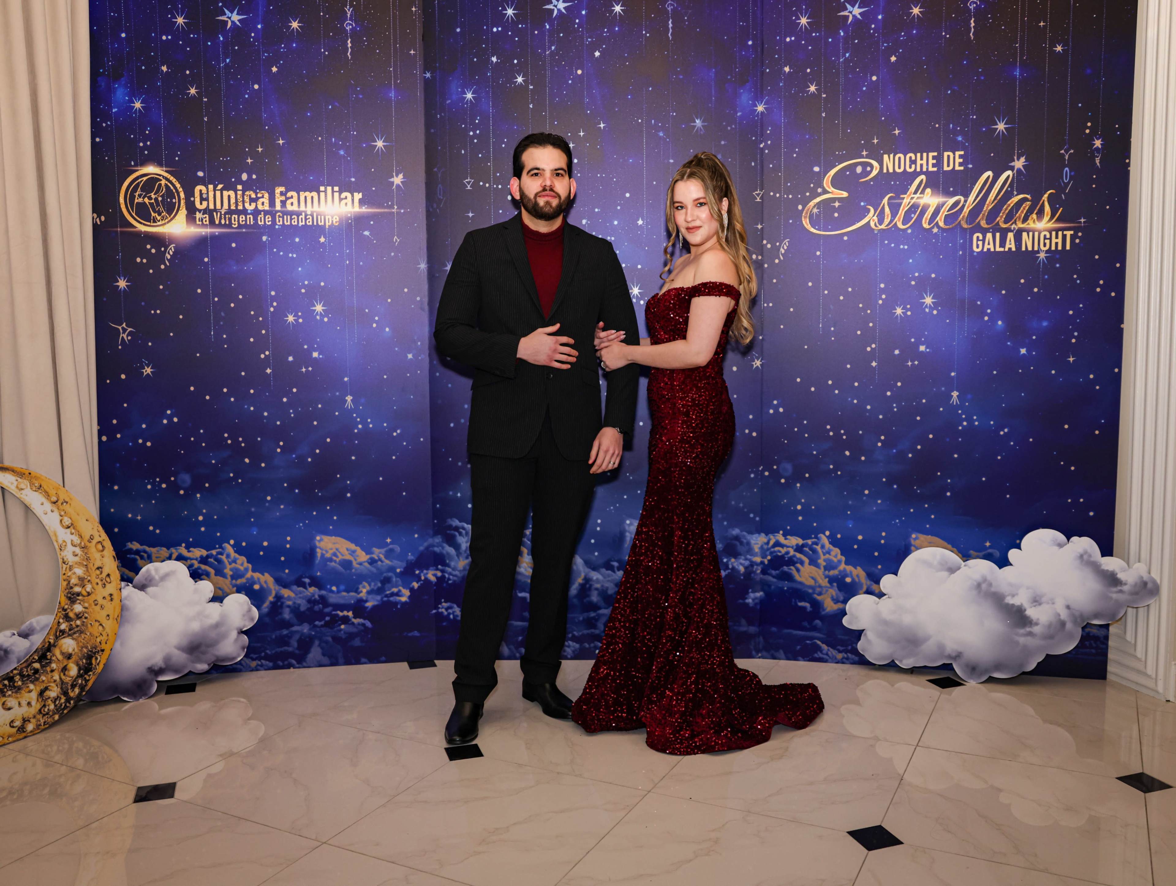 A man in a black suit and a woman in a red sequined gown pose together in front of a starry backdrop at an event called "Noche de Estrellas Gala Night."