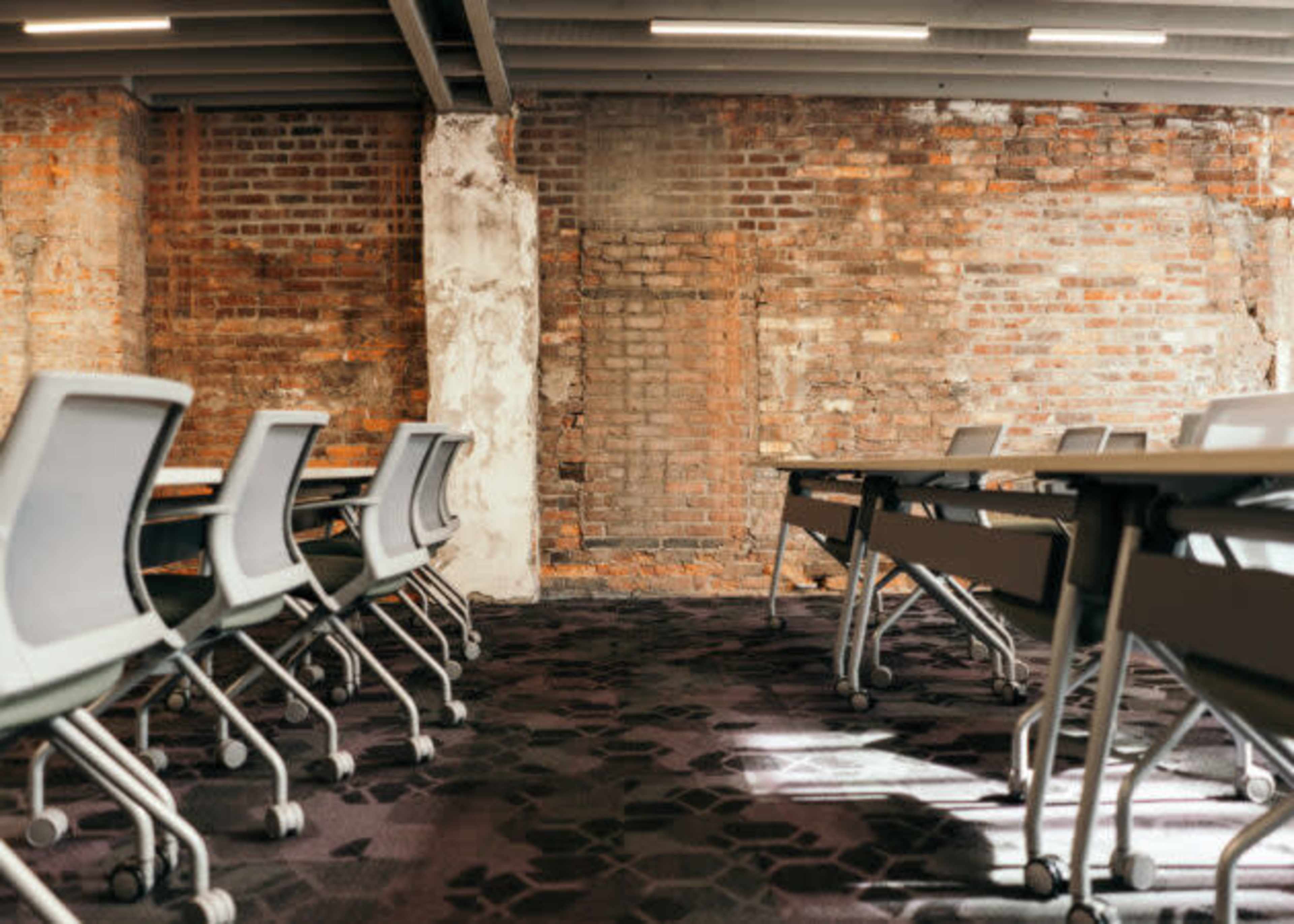 The image shows a conference room with modern chairs facing a row of tables against a brick wall background.