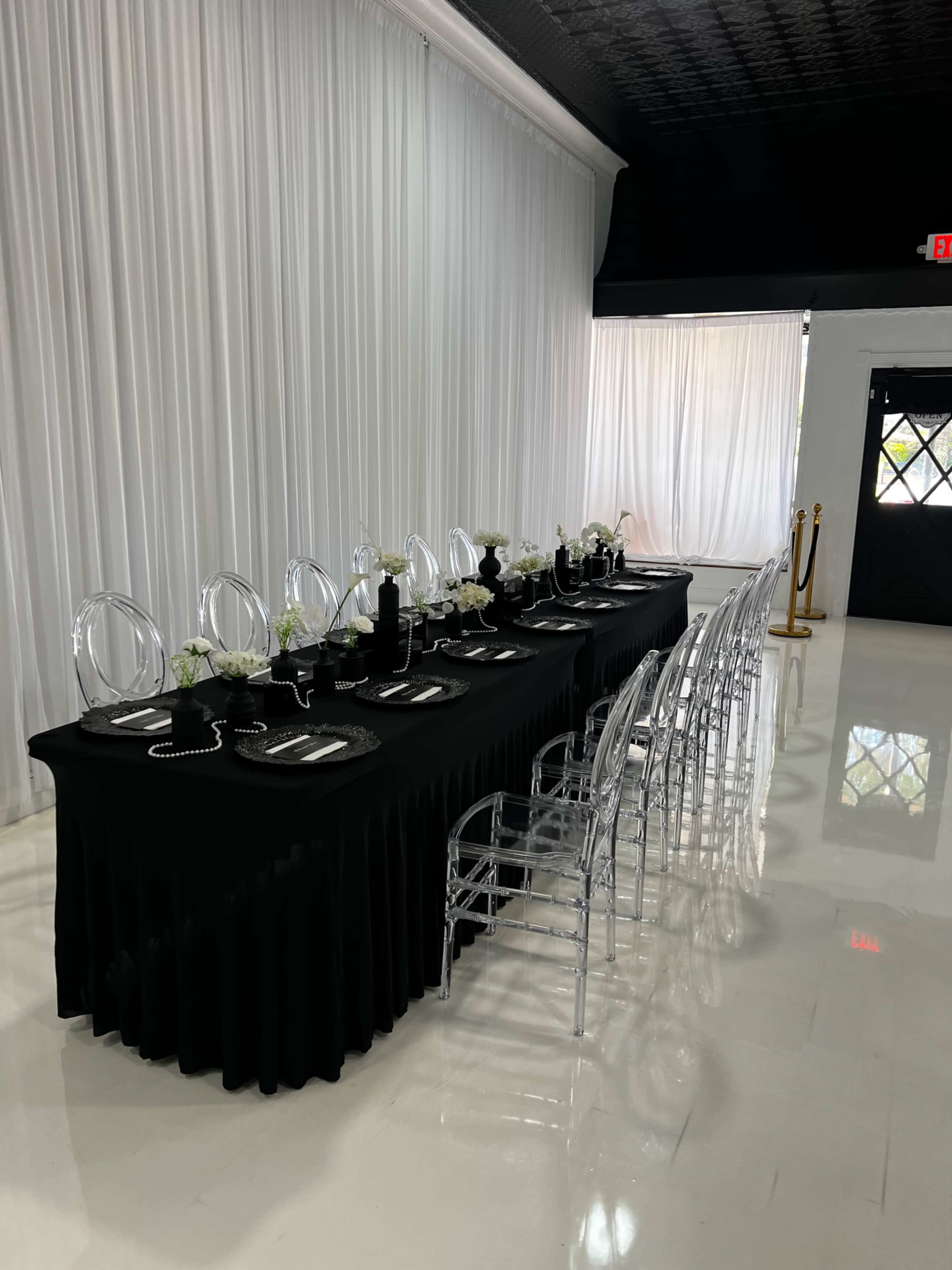 A long, black table is set for a formal dining event, adorned with white flowers and clear, ghost chairs in a well-lit space.