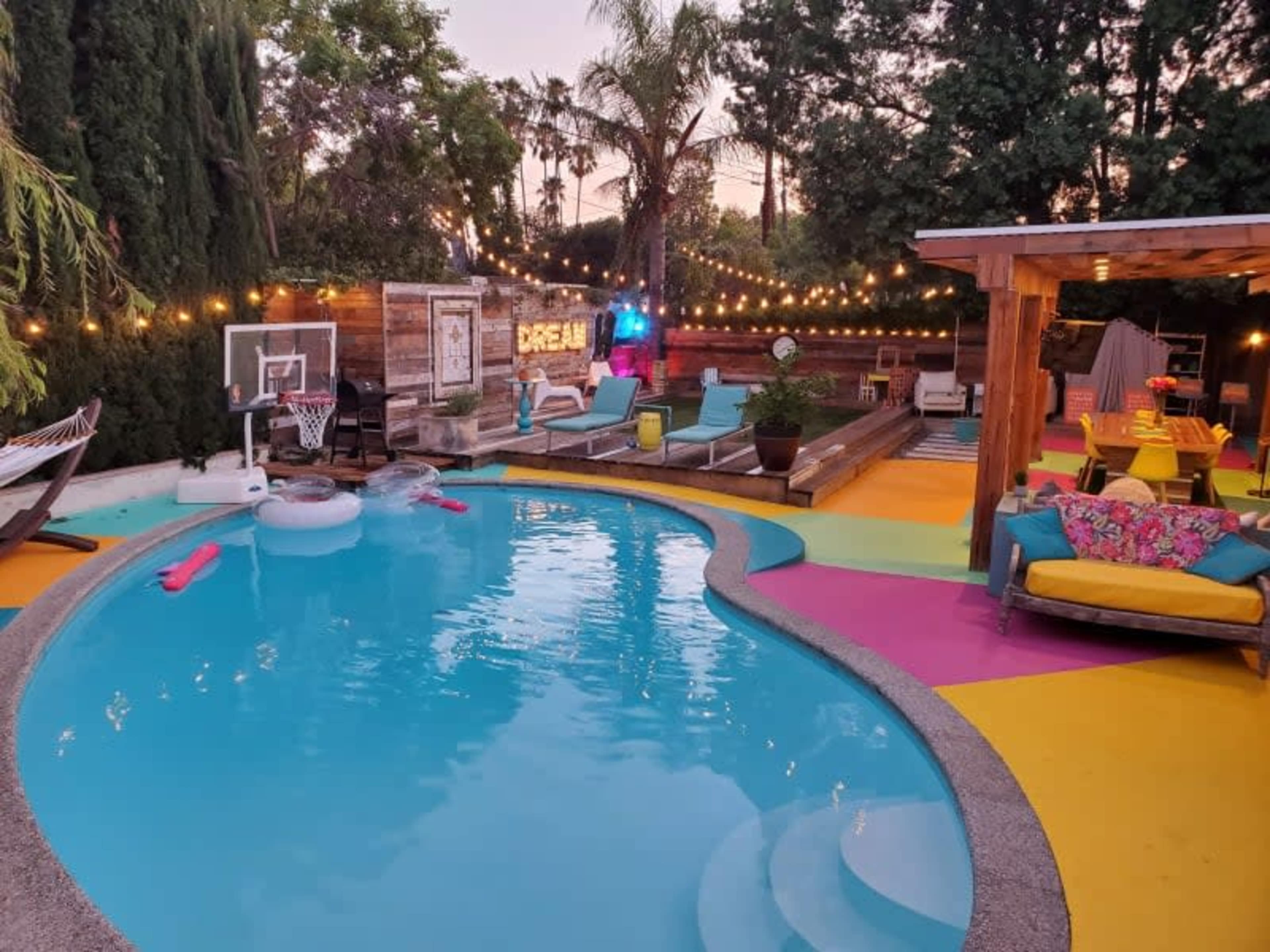 The image shows a colorful backyard featuring a swimming pool, lounge chairs, and string lights illuminating the area.