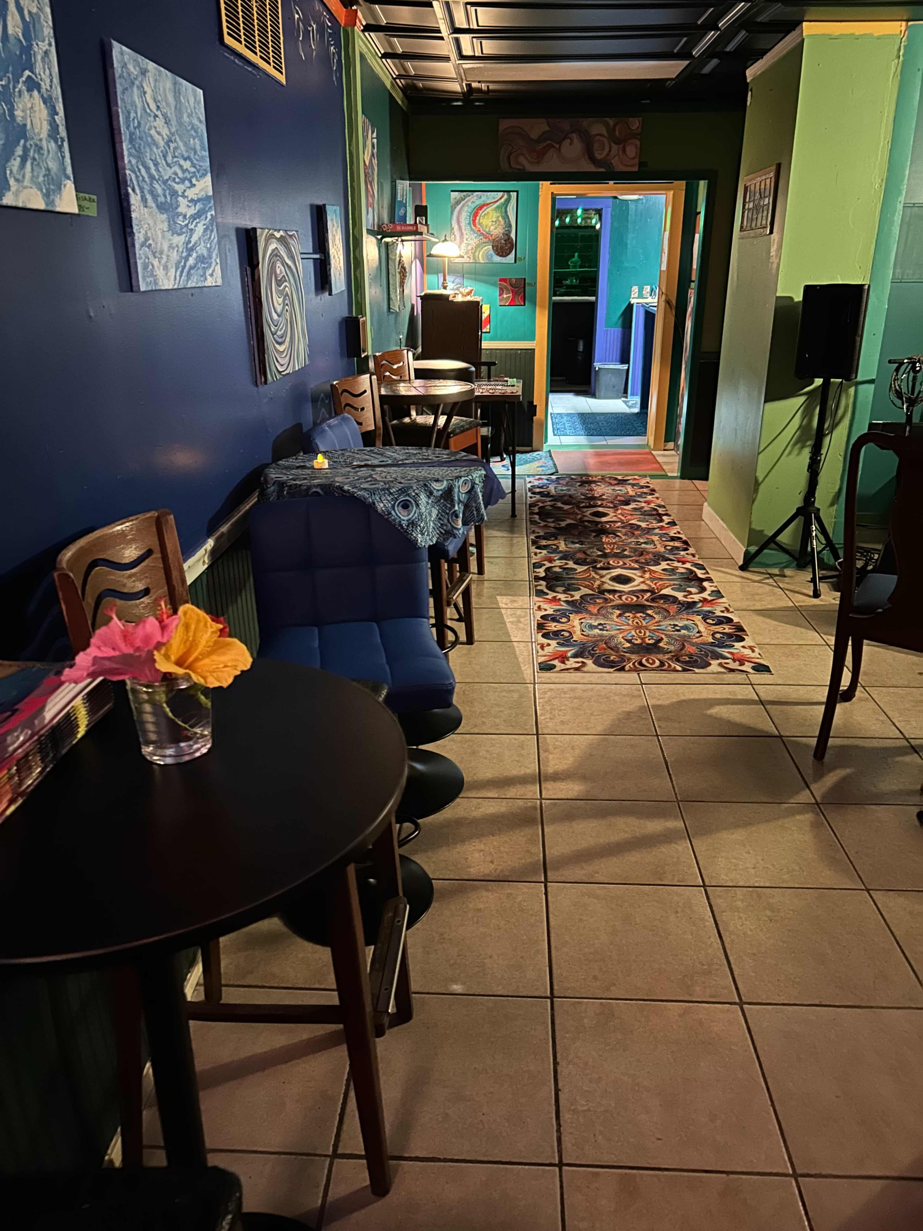 The image shows a cozy café interior with colorful walls, a patterned rug, and seating along the sides of a narrow pathway leading to the entrance.
