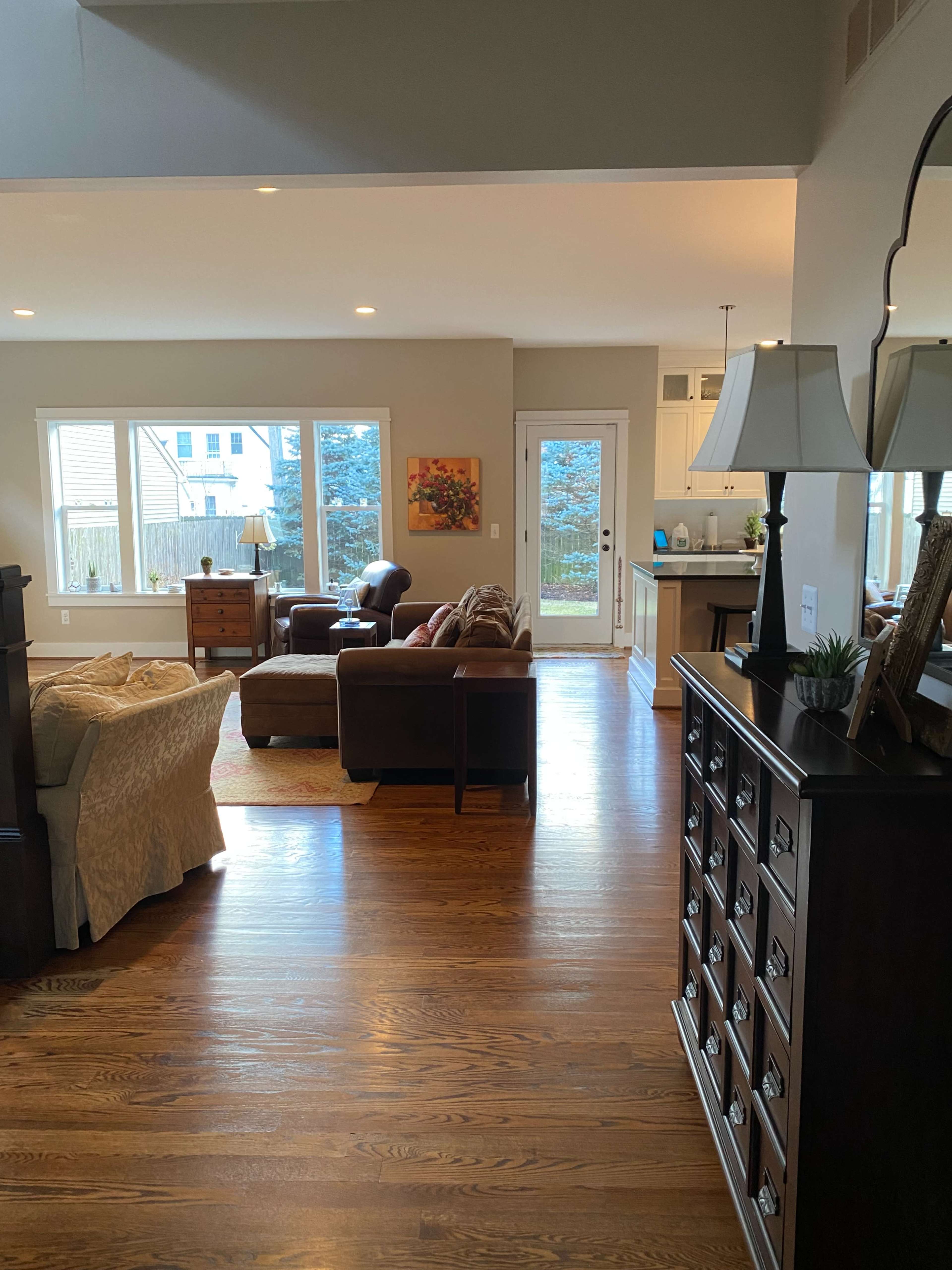 The image shows a living room area with large windows, hardwood flooring, and furniture including a sofa, chairs, and a wooden cabinet.