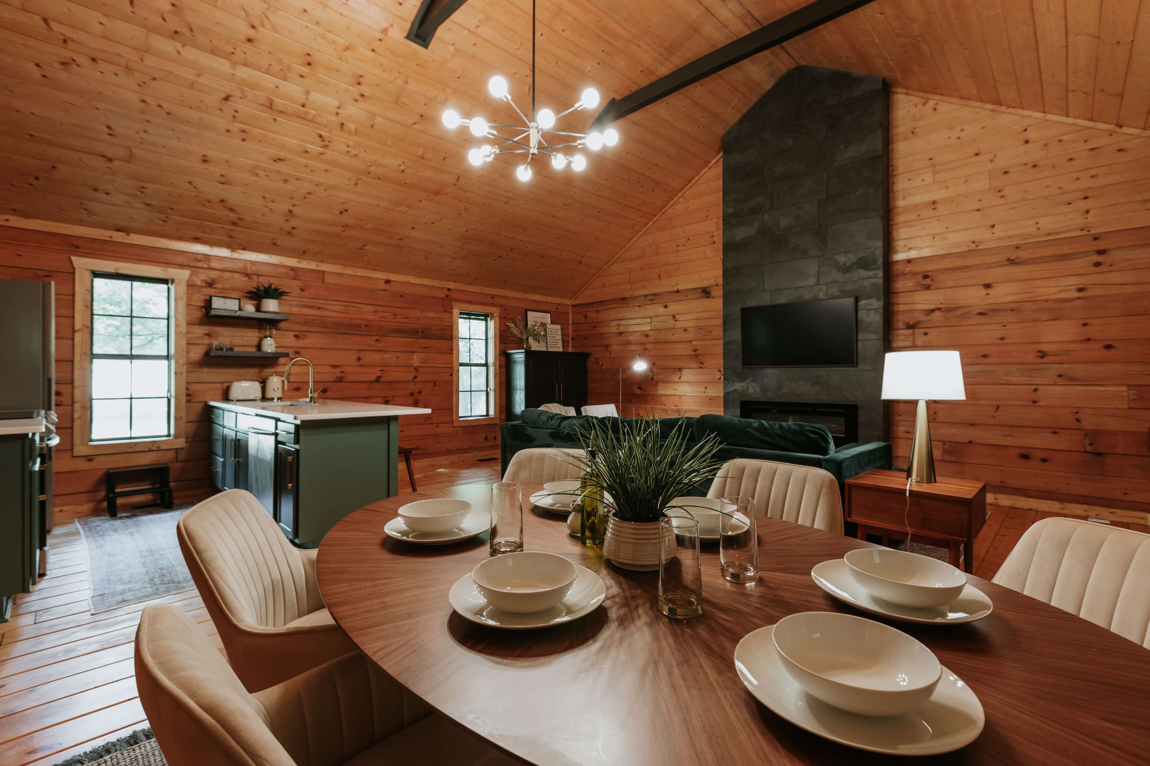 A cozy cabin interior features a dining table set for six, a modern kitchen area, and a living space with a large sofa and a wall-mounted TV.