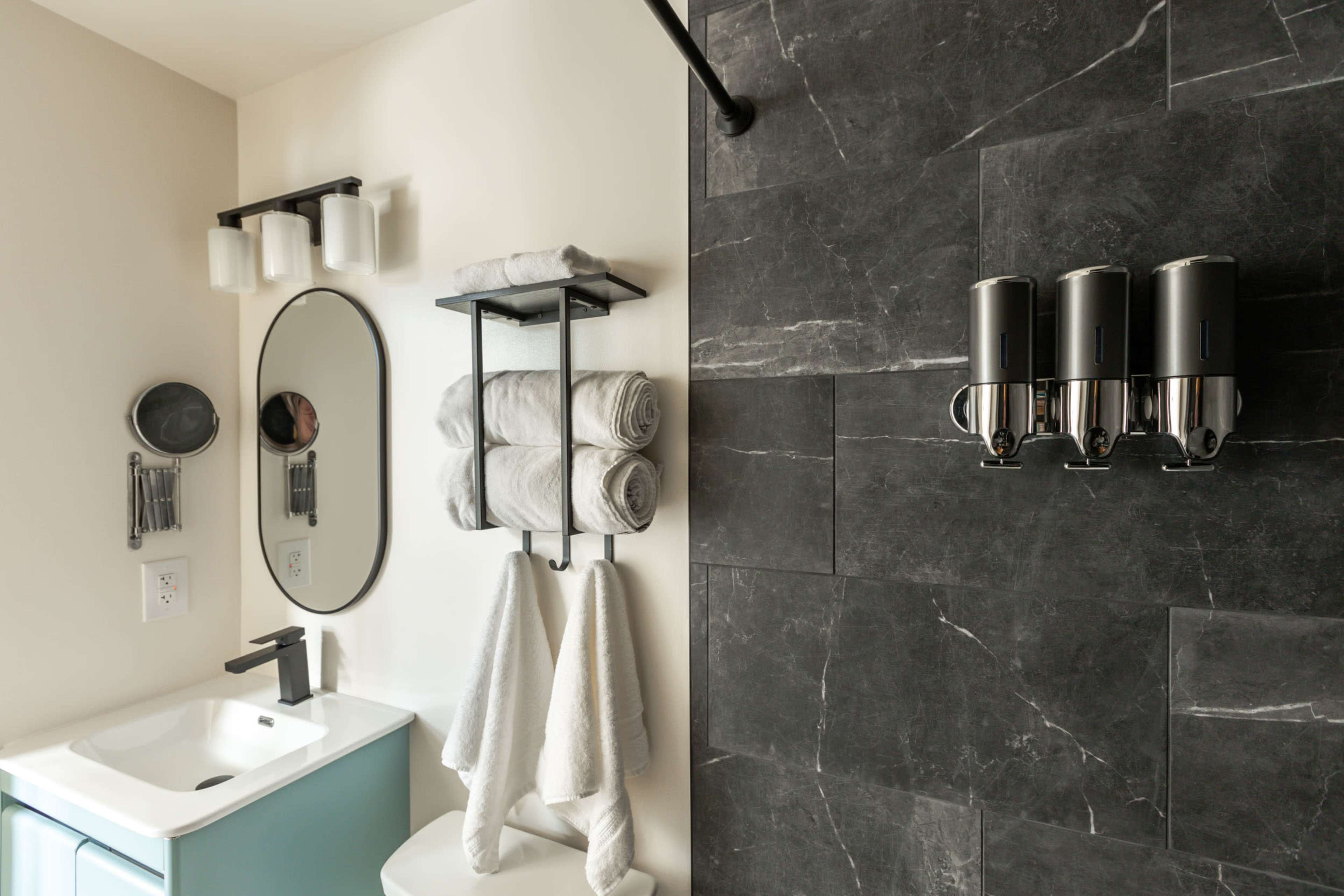 The image shows a modern bathroom featuring a mirror, a sink with a cabinet, neatly stacked towels, and a wall-mounted dispenser for toiletries.