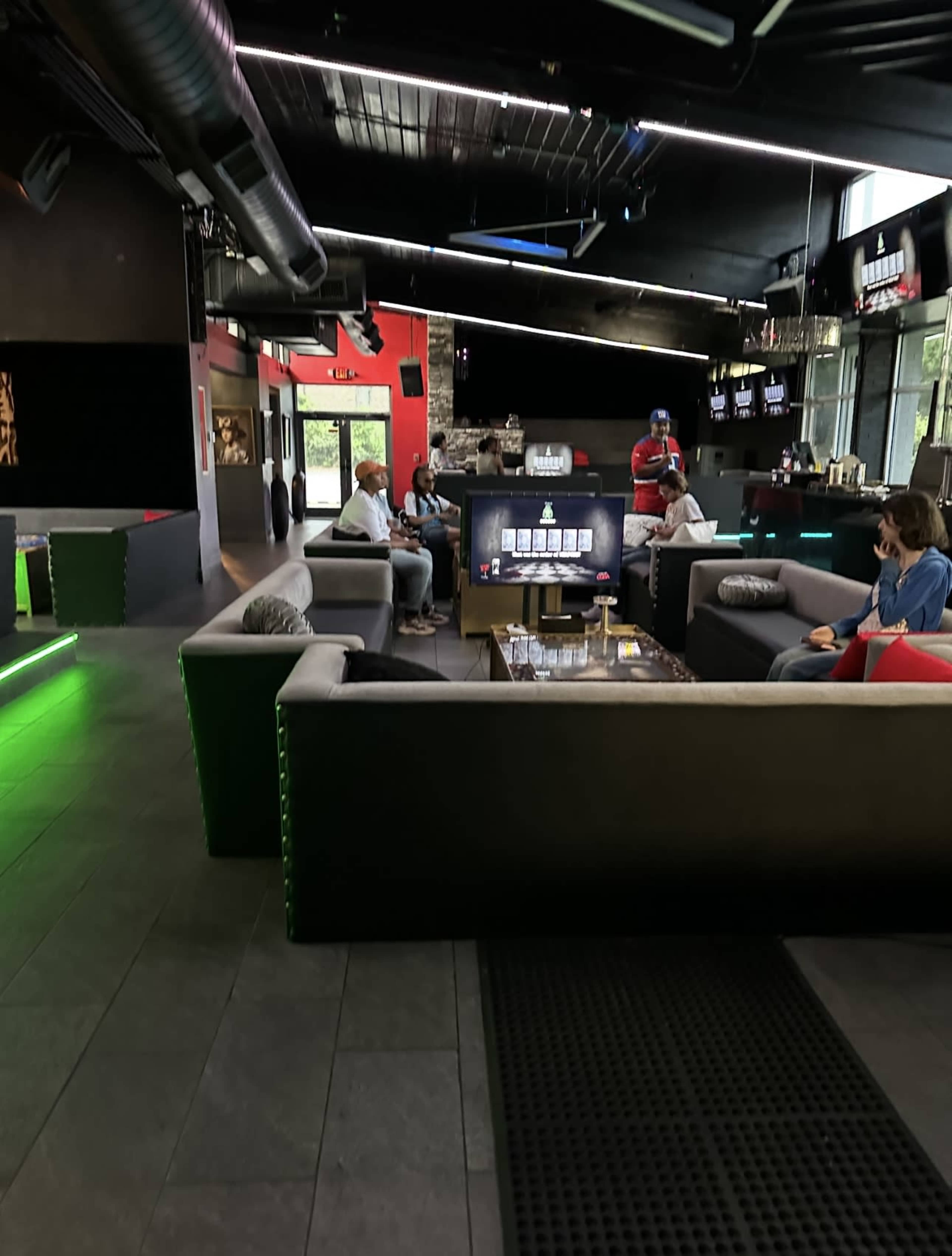 The image shows a modern lounge area with several people seated on large sofas around a coffee table, with a television visible in the center of the space.