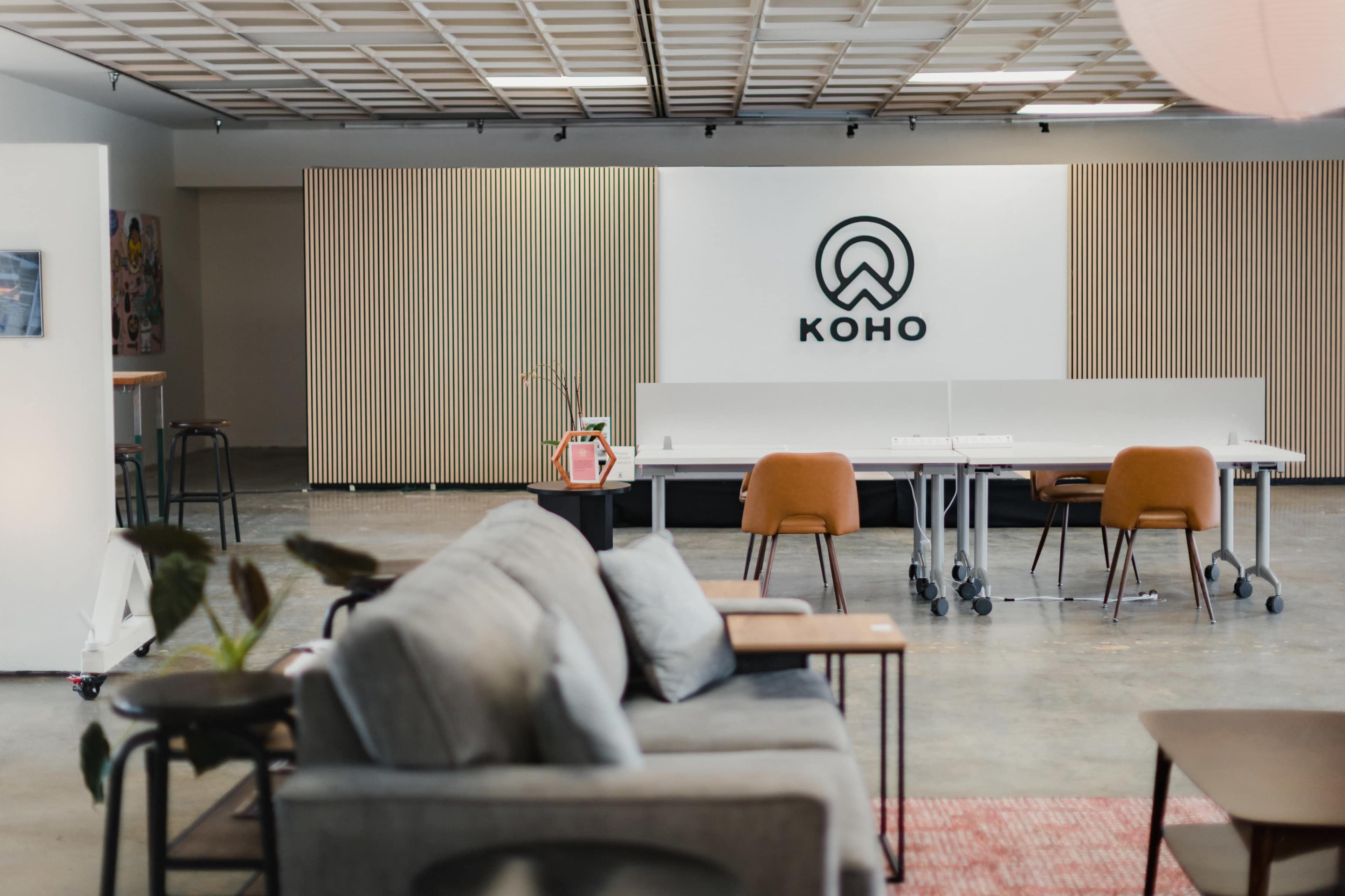 The image shows a modern office space with a logo for "KOHO" on the wall, featuring a seating area and a row of white desks with chairs.