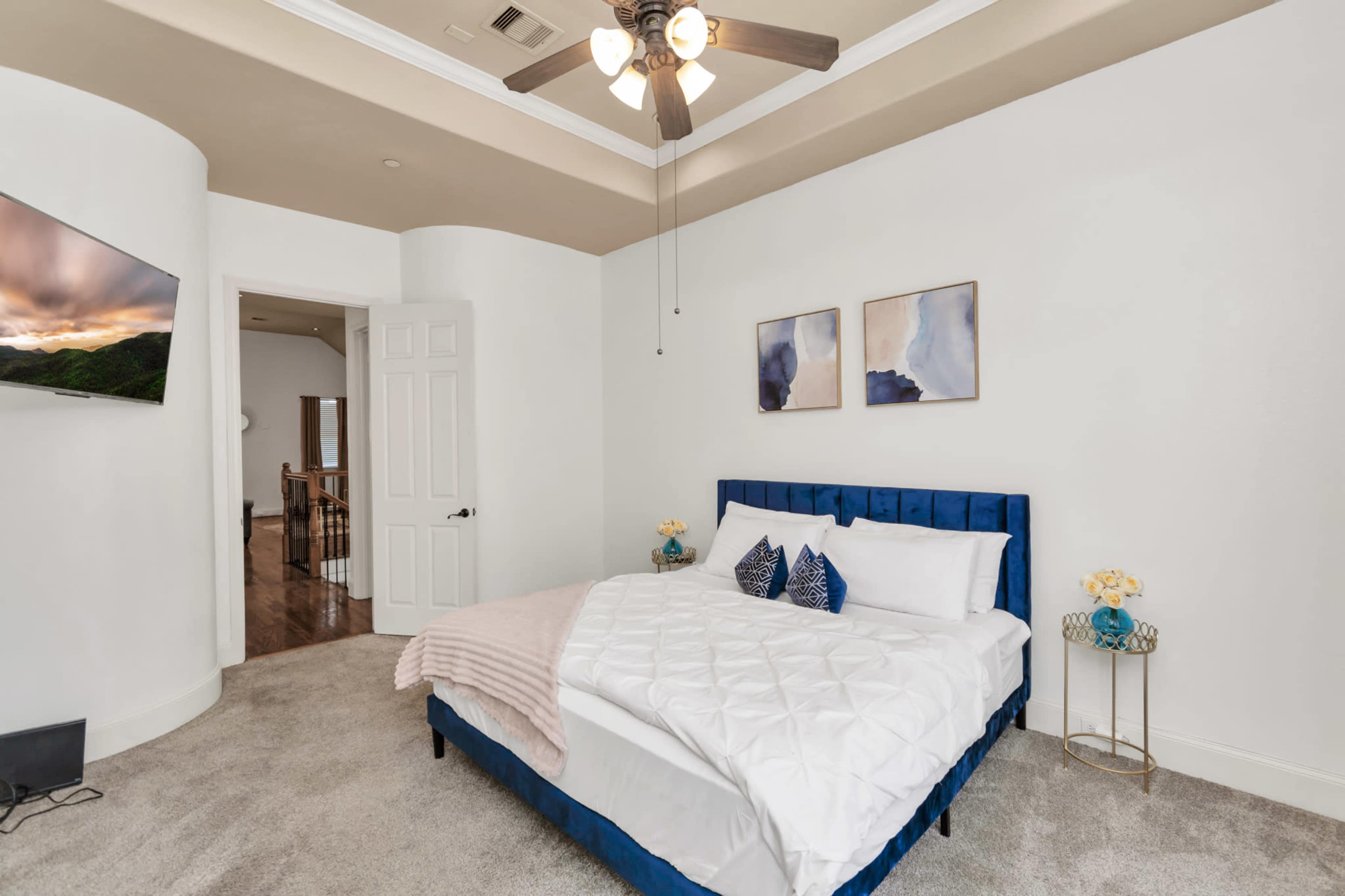 A well-lit bedroom featuring a blue upholstered bed with decorative pillows and artwork on the walls, alongside a ceiling fan and a doorway leading to another room.