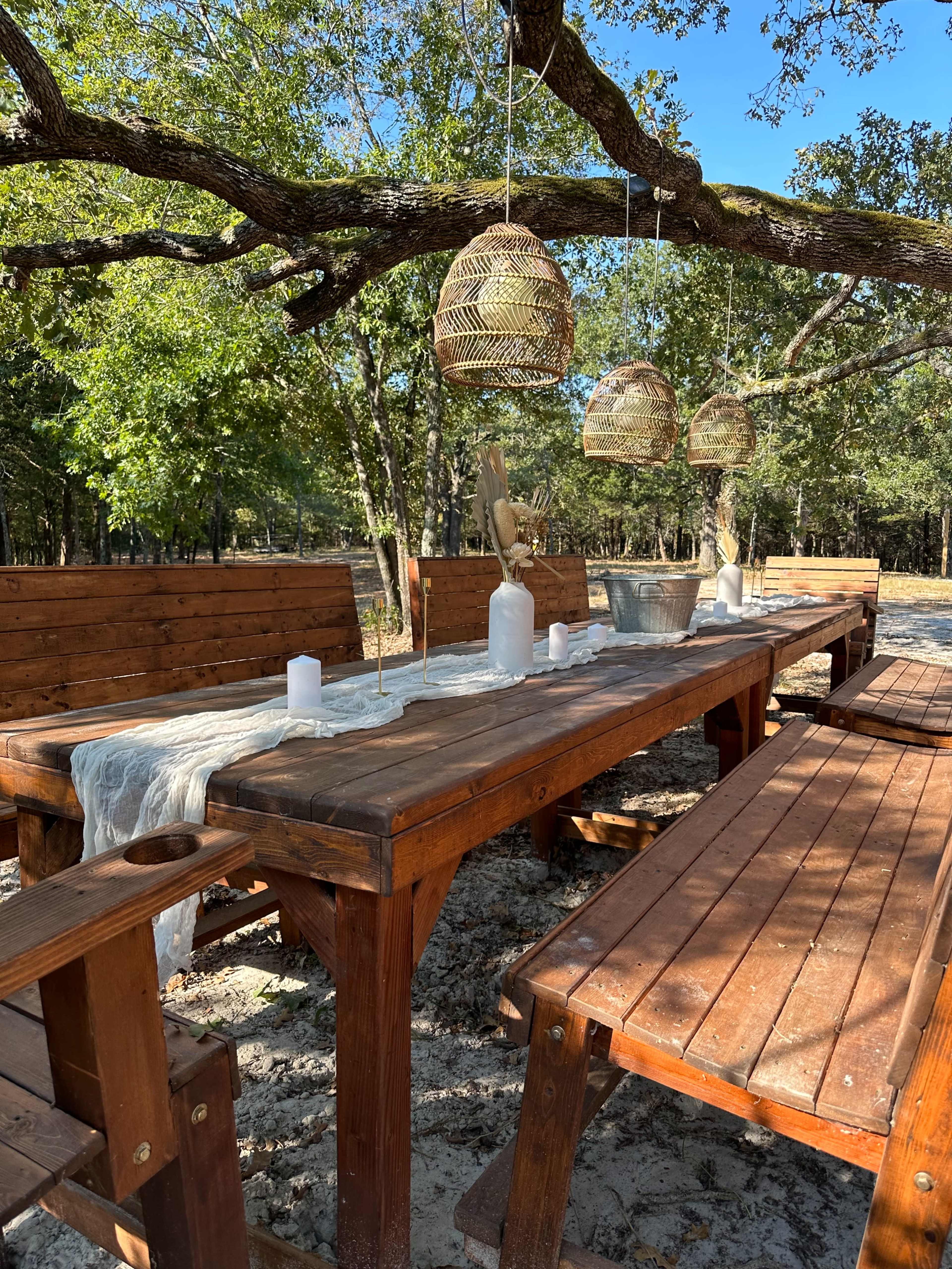 A wooden outdoor dining table is set with decorative items and candles under hanging light fixtures, surrounded by chairs in a forested area.