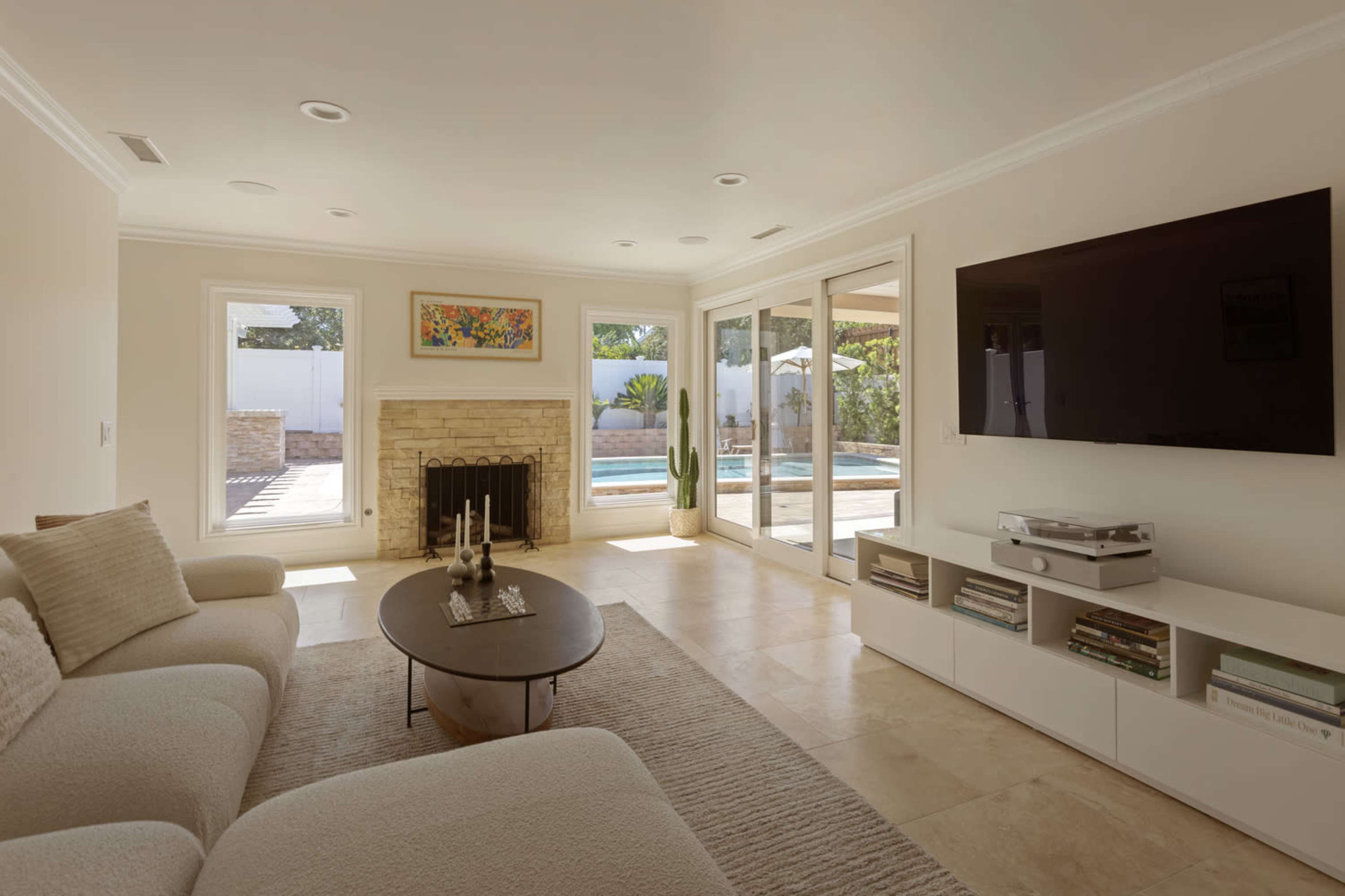 A modern living room features a light-colored sectional sofa, a round coffee table, a stone fireplace, and large glass doors that open to a pool area.