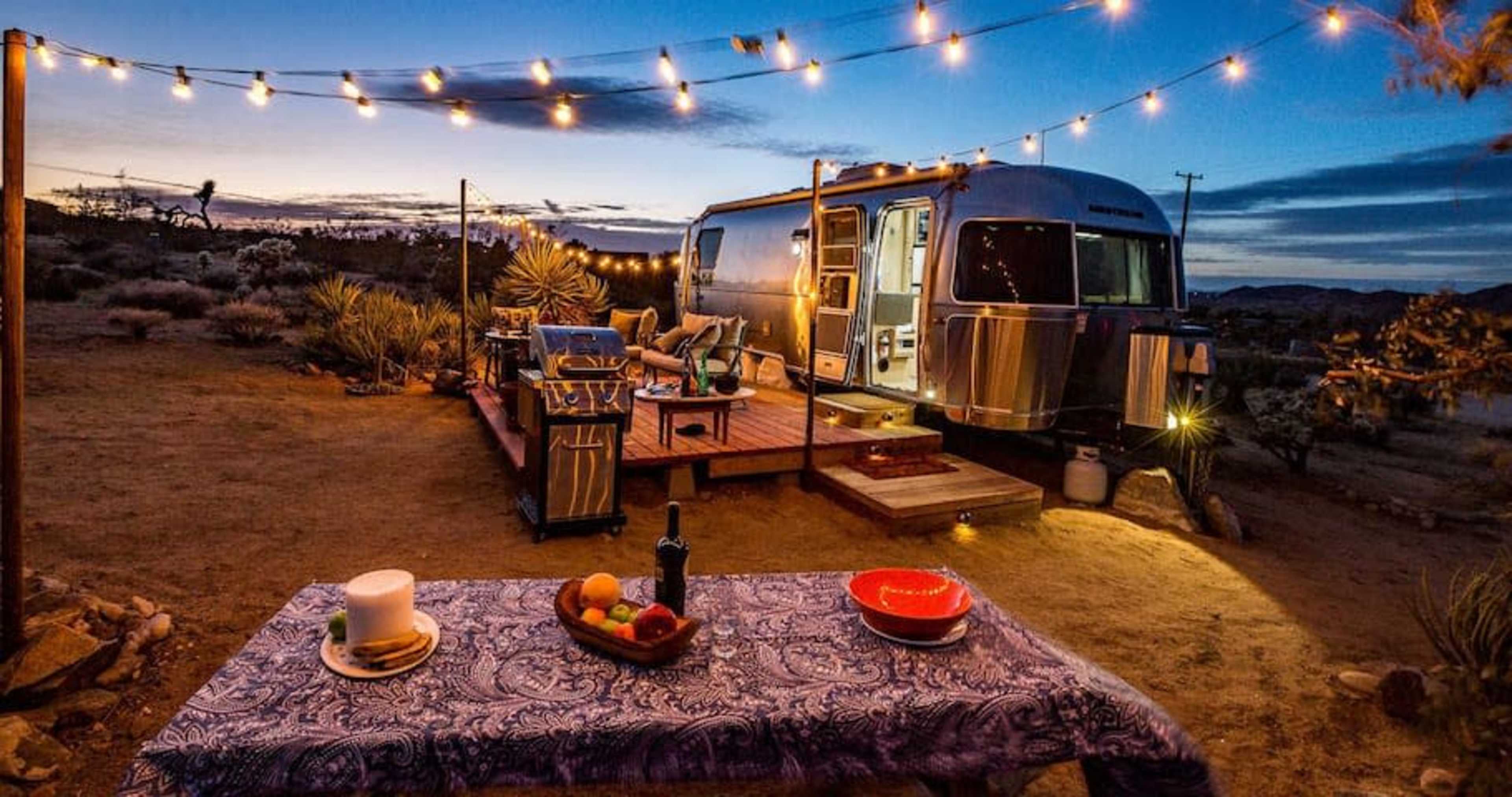 A retro Airstream trailer is parked on a desert lot, illuminated by string lights and surrounded by outdoor furniture and a table set with fruit.