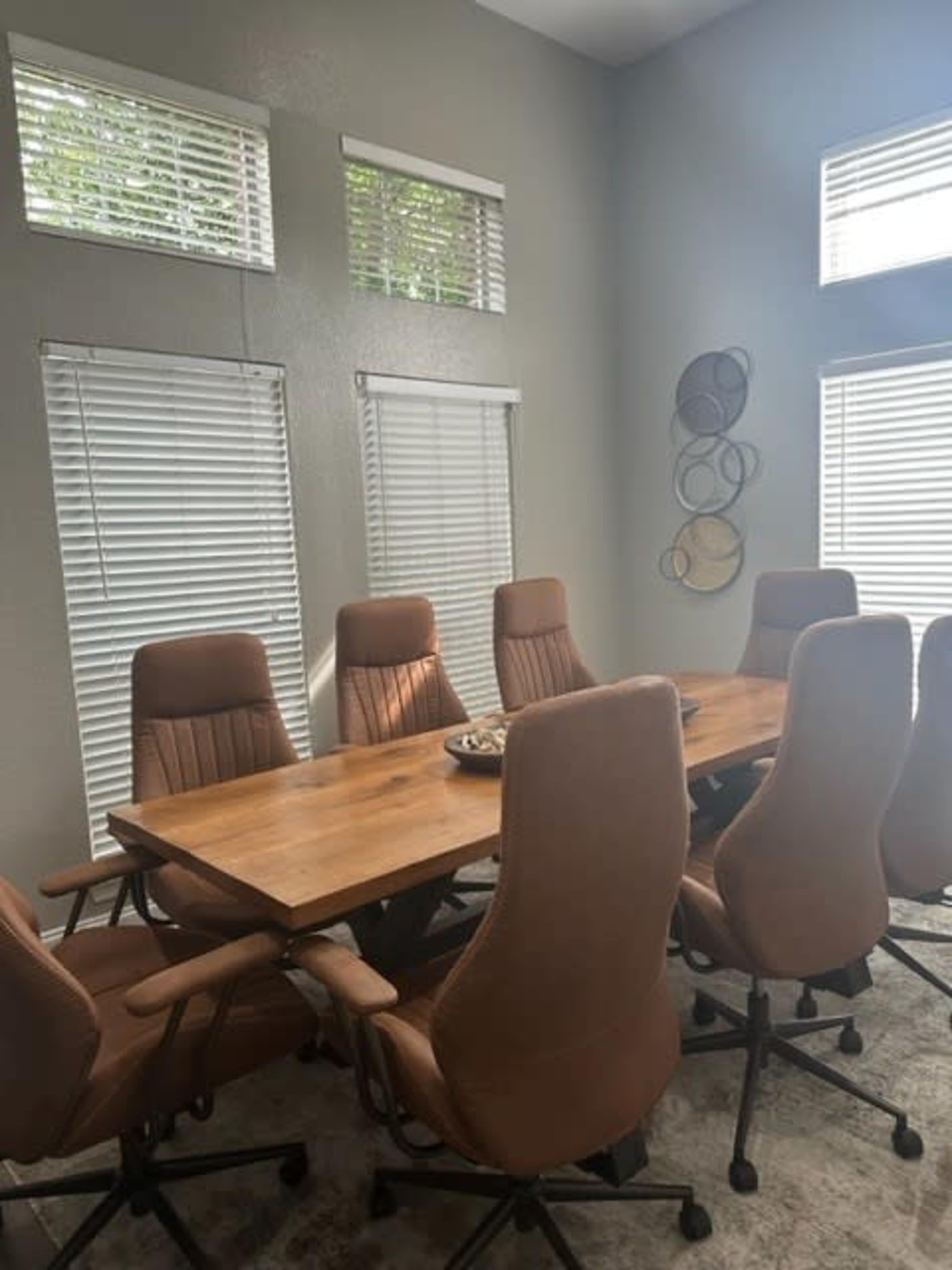 A modern conference room features a large wooden table surrounded by eight ergonomic office chairs, with sunlight streaming through slatted windows.