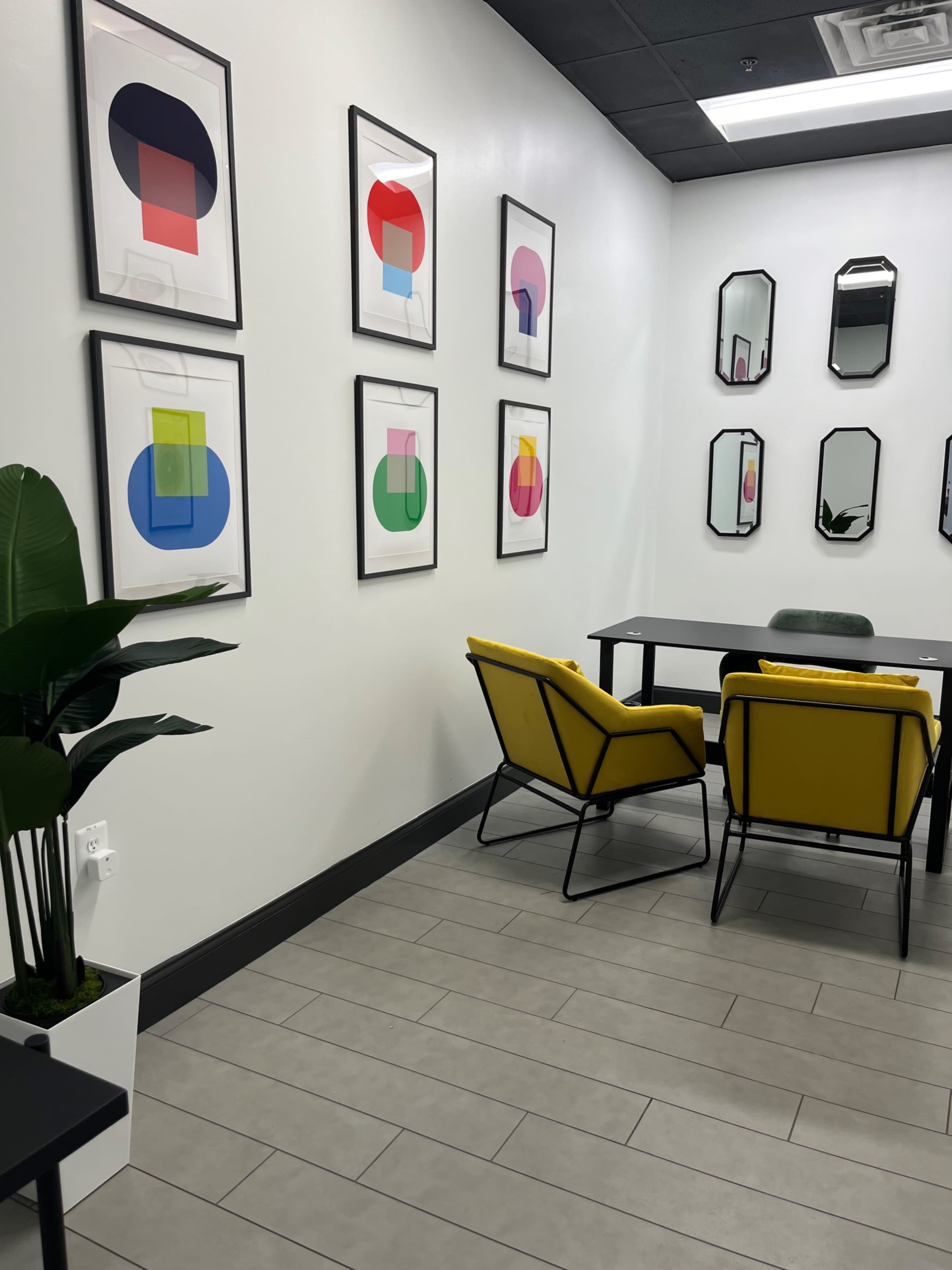 The image shows a modern room with yellow chairs, a rectangular table, and colorful framed artwork on the walls, alongside several mirrors.