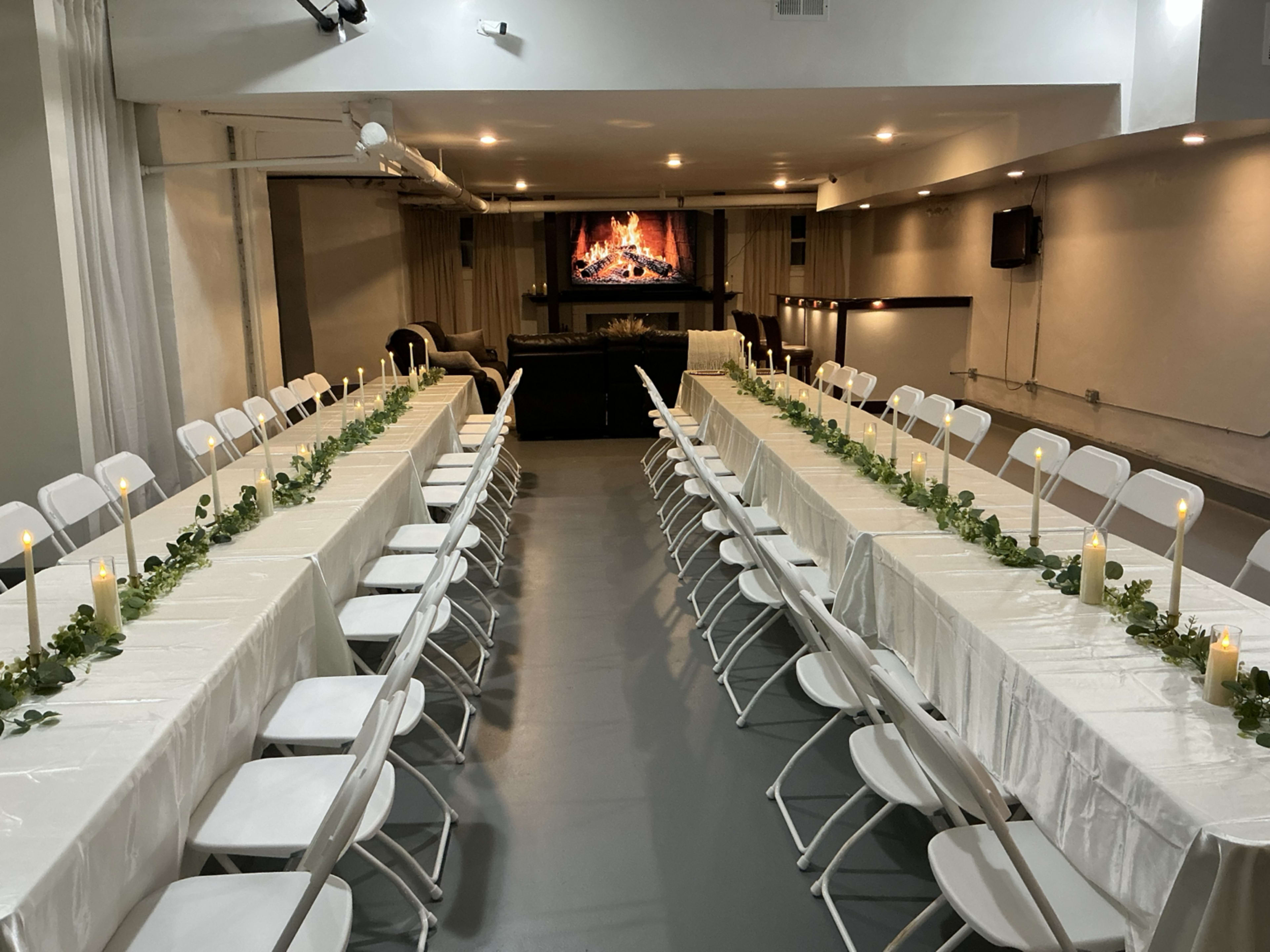 A spacious dining area is set up with long tables covered in white tablecloths, arranged in parallel lines and adorned with greenery and candles, with a fireplace display visible on a screen at the far end.