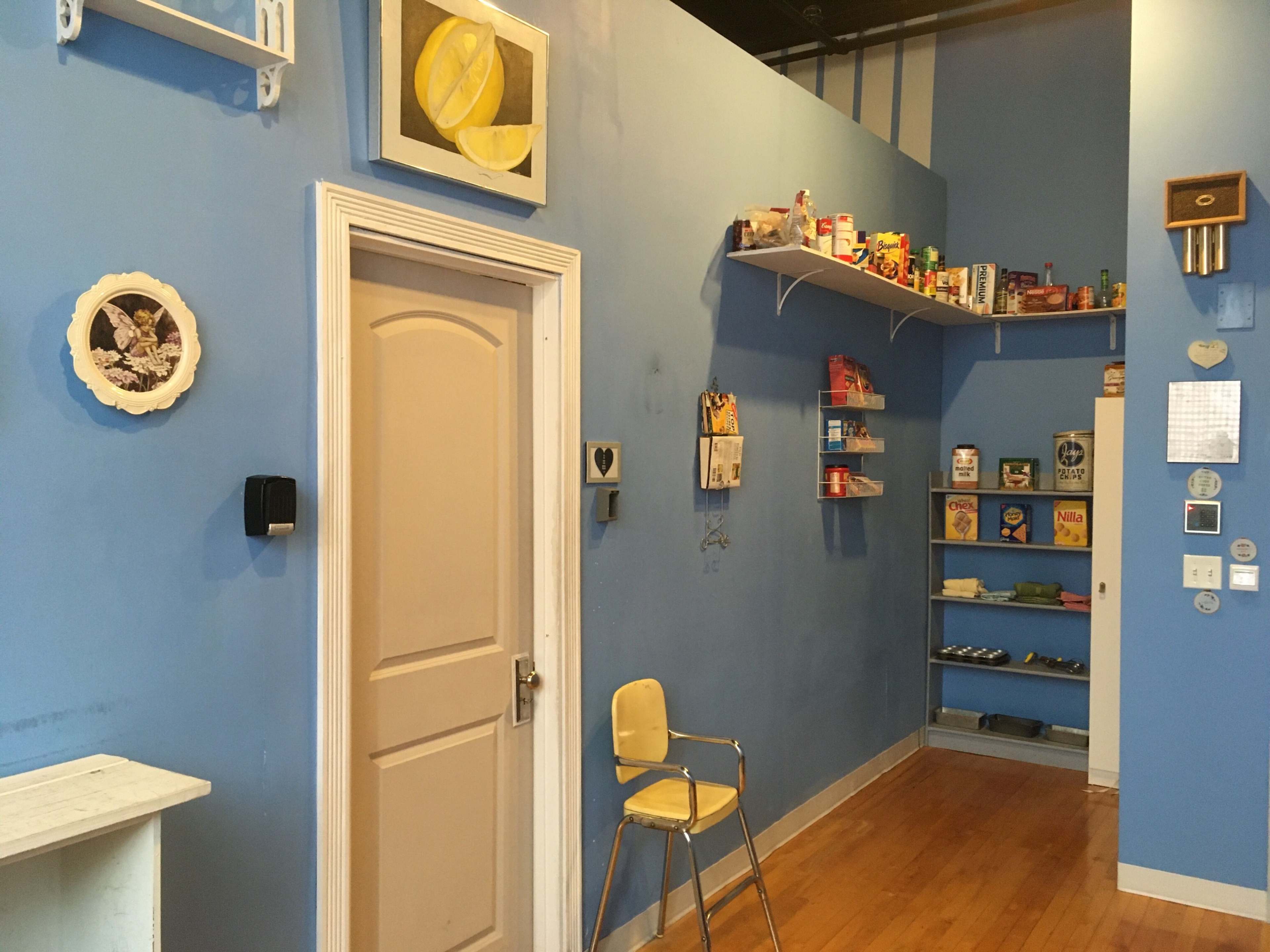 A blue-walled room with a door, a chair, and shelves displaying various food items and decorative pieces.