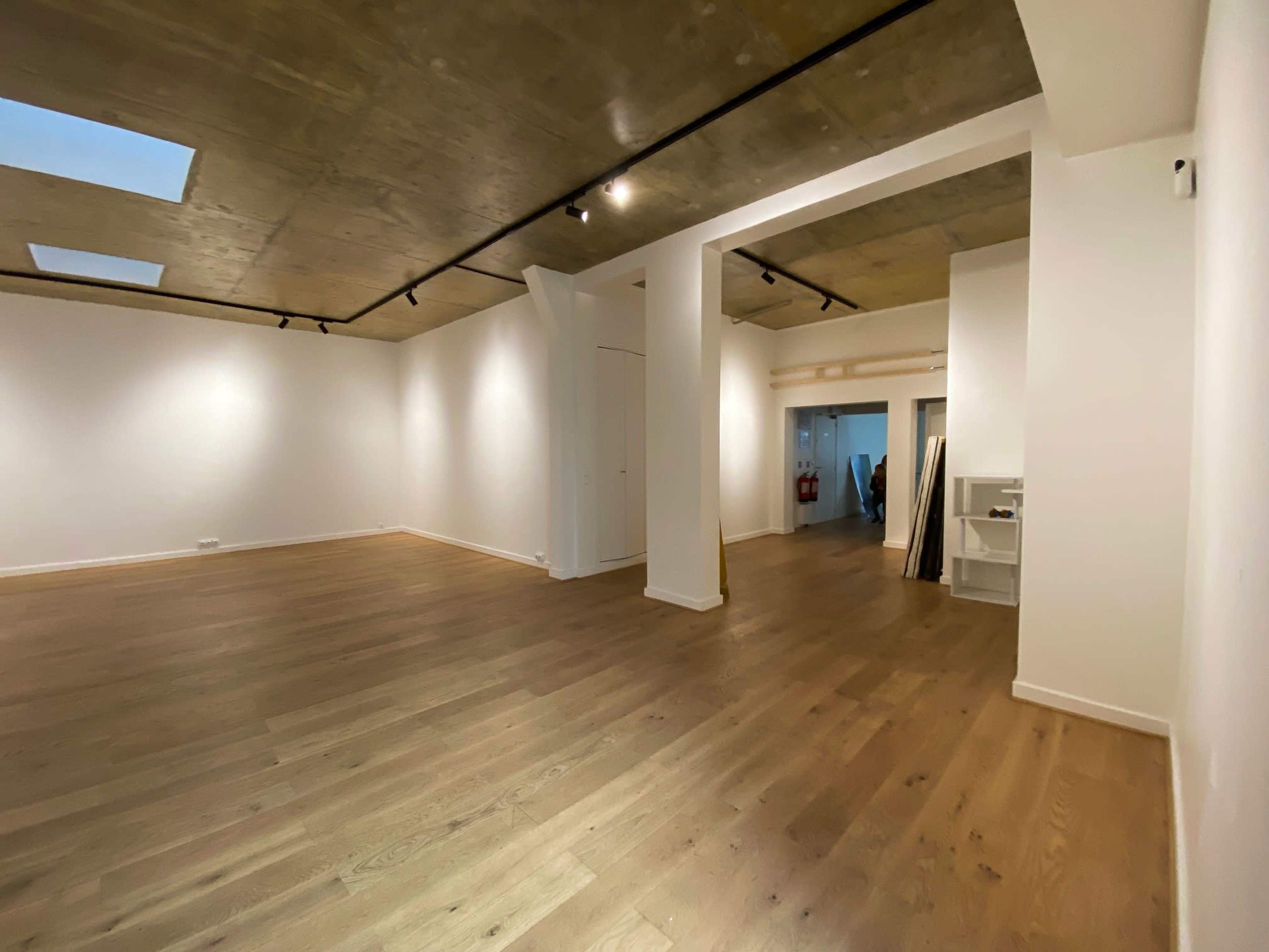 The image shows a spacious, empty room with wooden flooring and white walls, featuring a ceiling with skylights and recessed lighting.