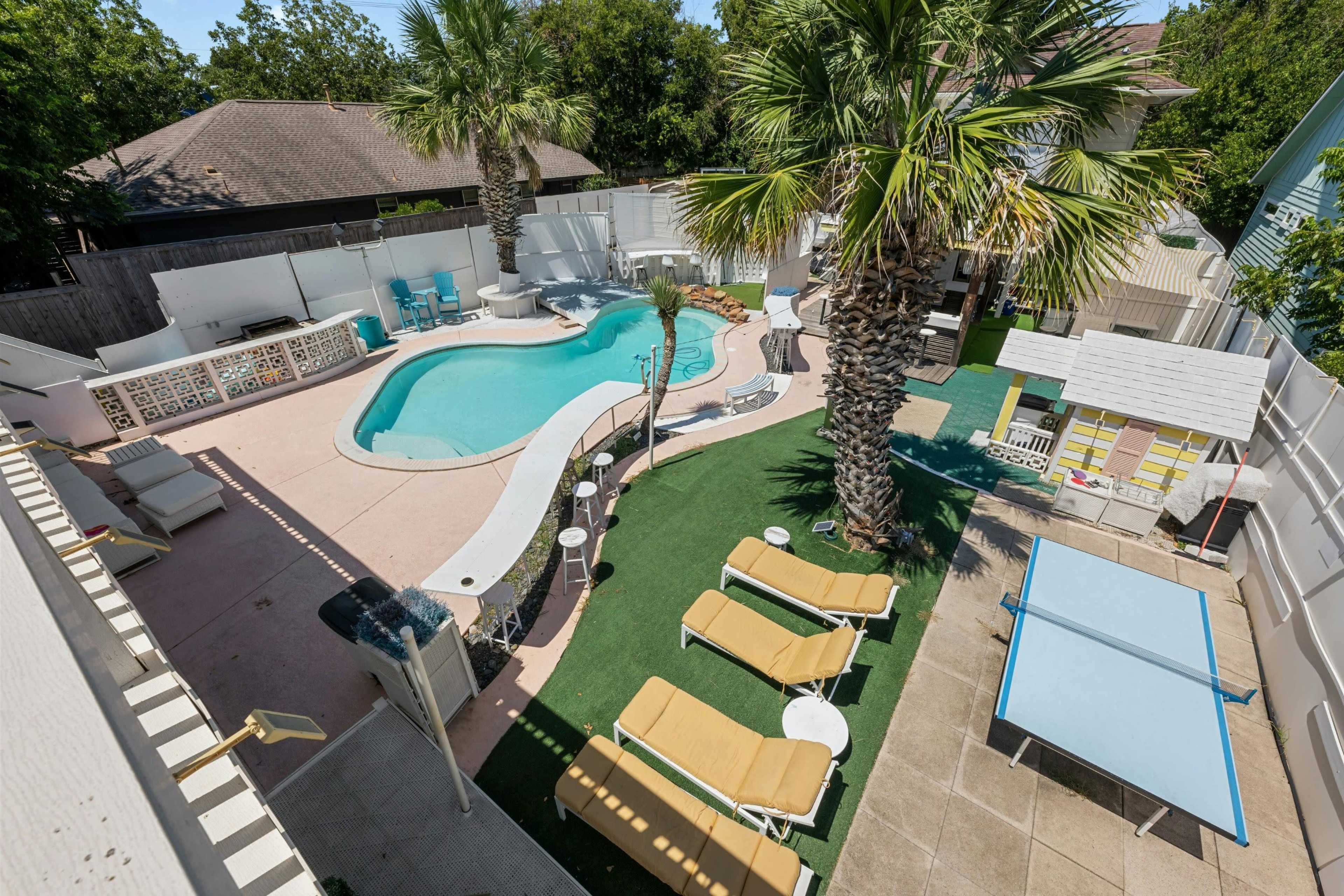 The image shows a backyard featuring a swimming pool, lounge chairs, palm trees, and a small playhouse, with a ping pong table on the patio.