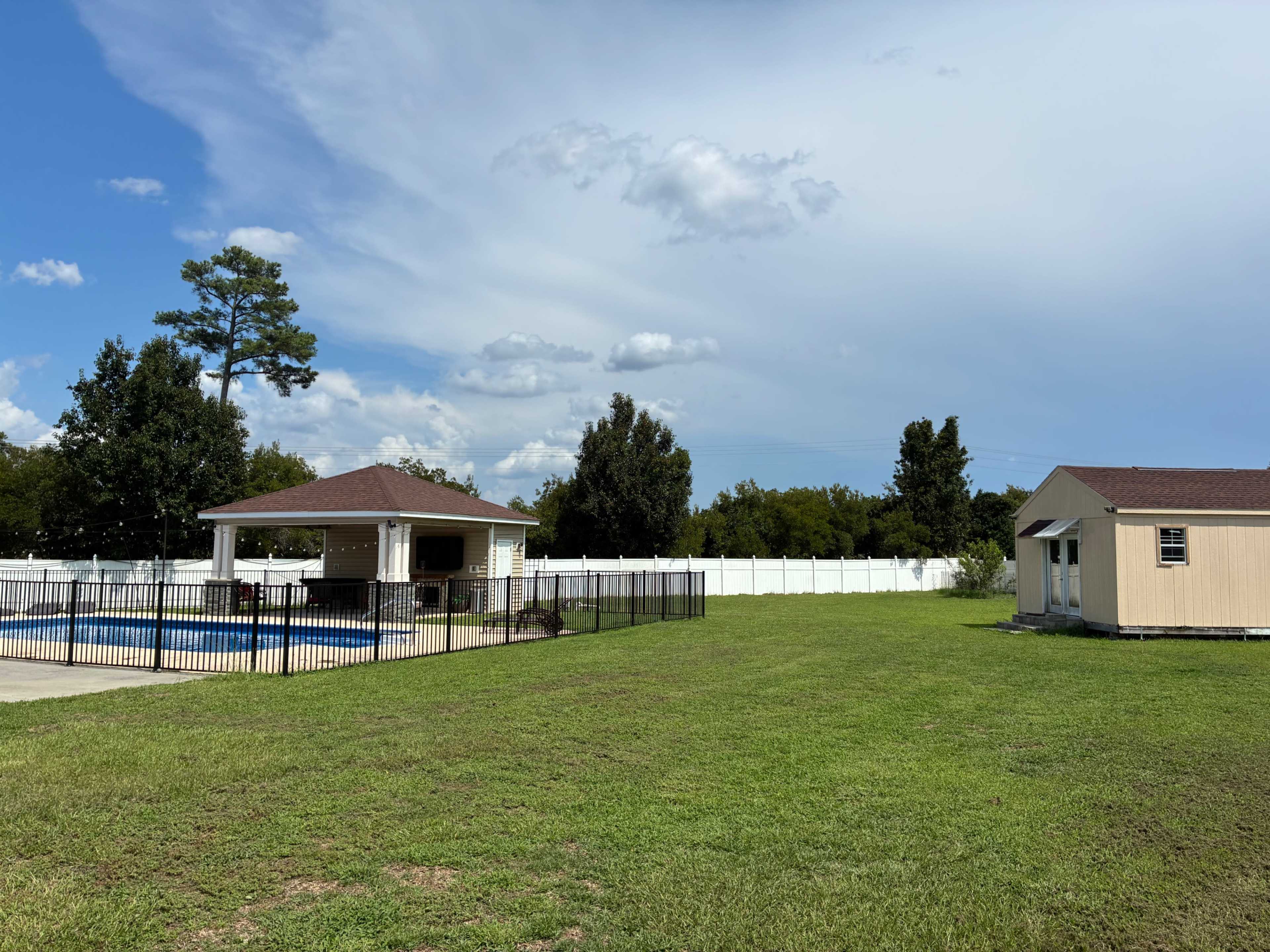 The image shows a grassy area with a fenced swimming pool and a nearby gazebo on the left, and a small building on the right surrounded by trees and a blue sky.