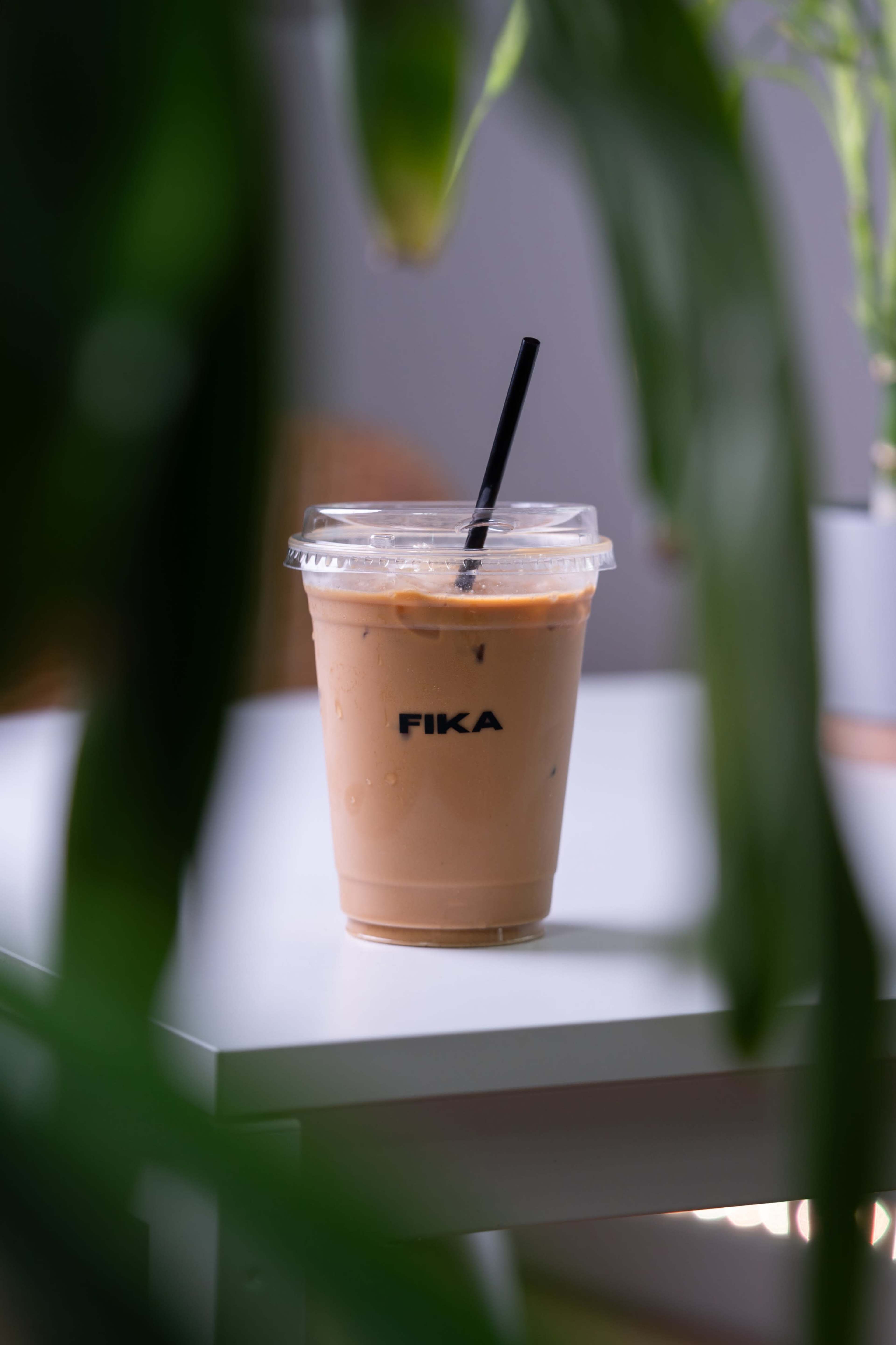 A clear plastic cup of iced coffee with a black straw sits on a table, partially obscured by green plant leaves.