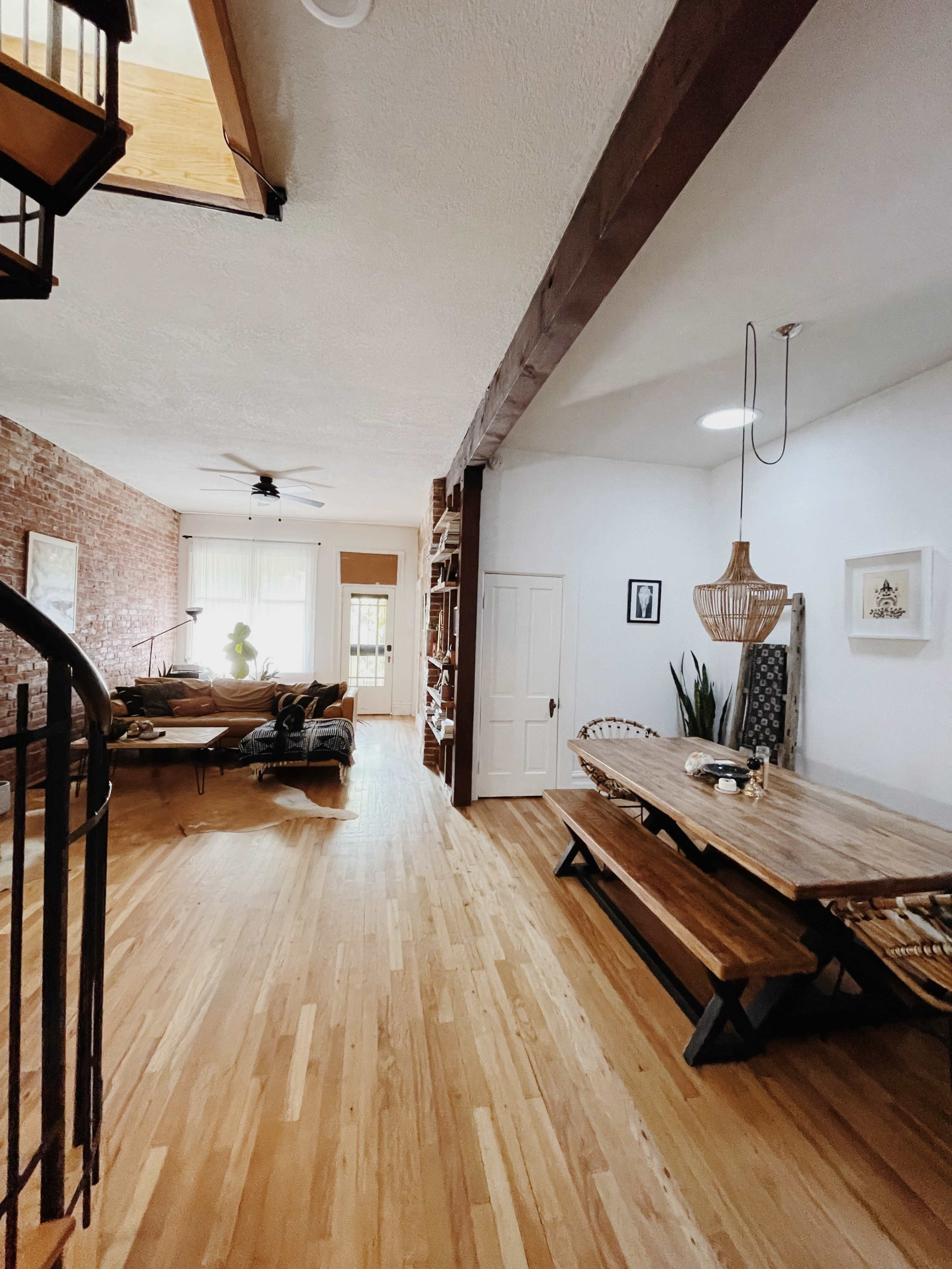 A spacious, open-concept living area with a wooden dining table, a brick accent wall, and a staircase leading to an upper level.