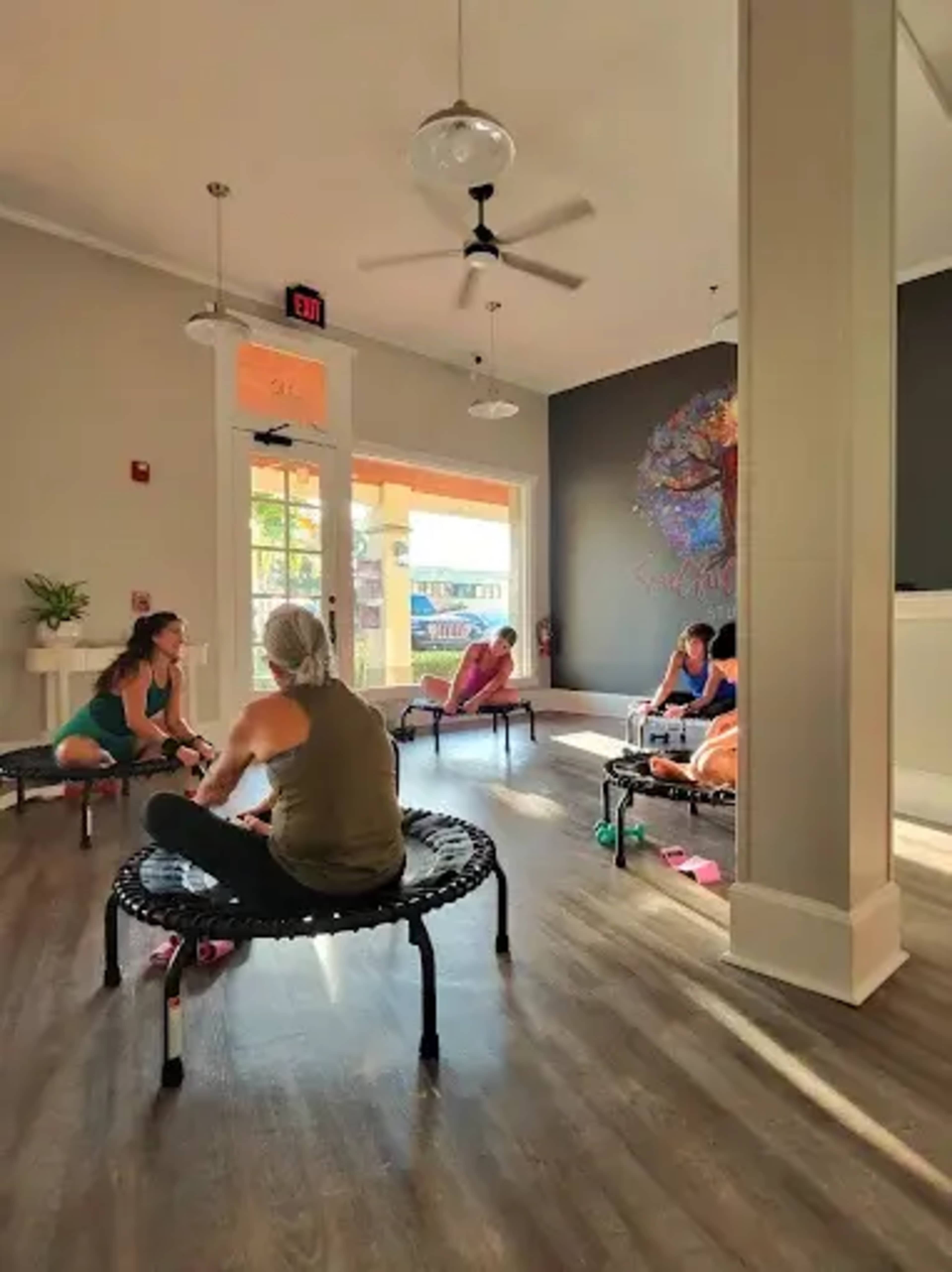 A group of people is seated on miniature trampolines in a bright, modern room with large windows and colorful wall art.