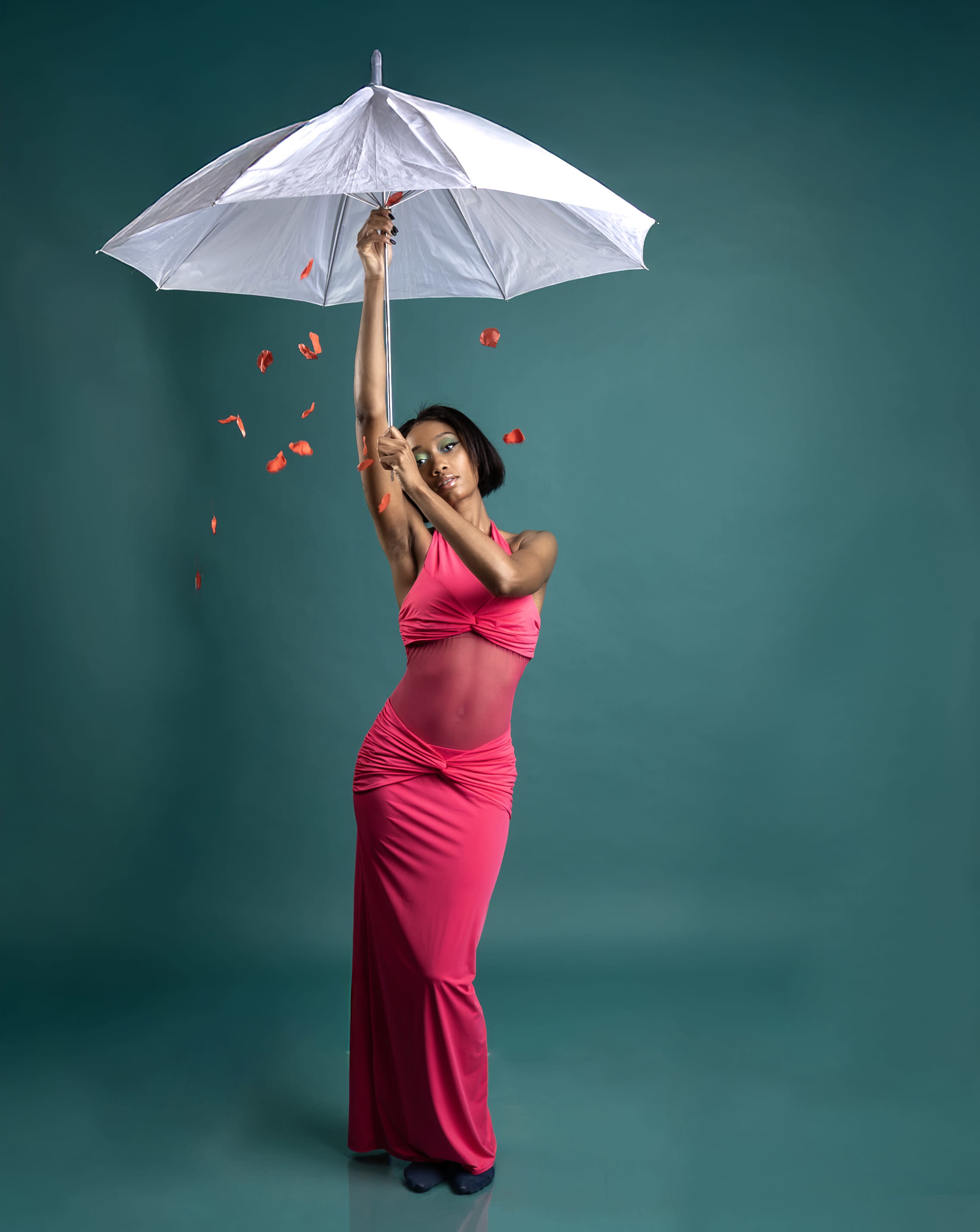 A person in a pink dress holds a white umbrella against a teal background, with rose petals cascading down.
