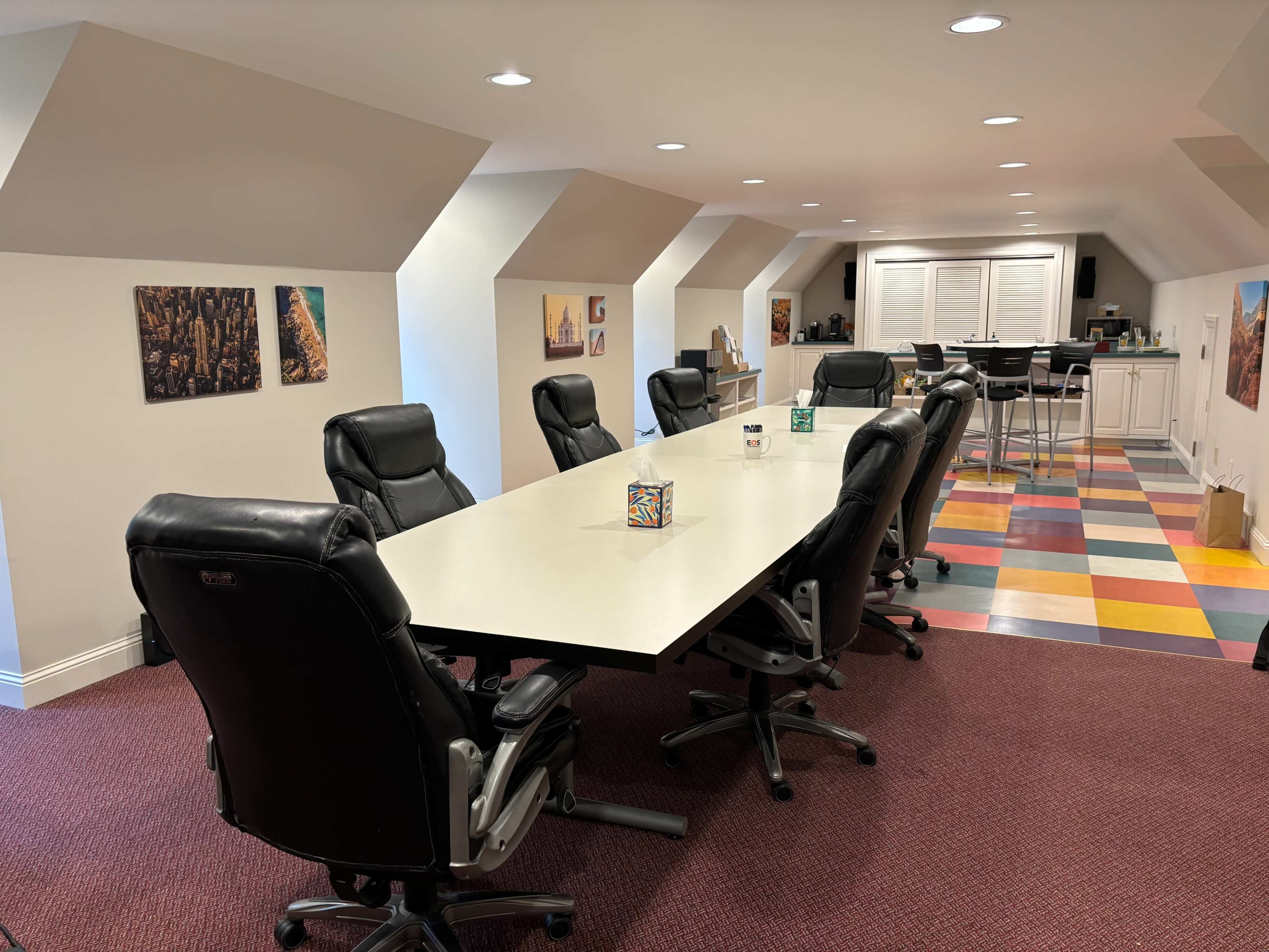 A large conference table surrounded by ergonomic black chairs is set up in a brightly colored meeting room with artwork on the walls.