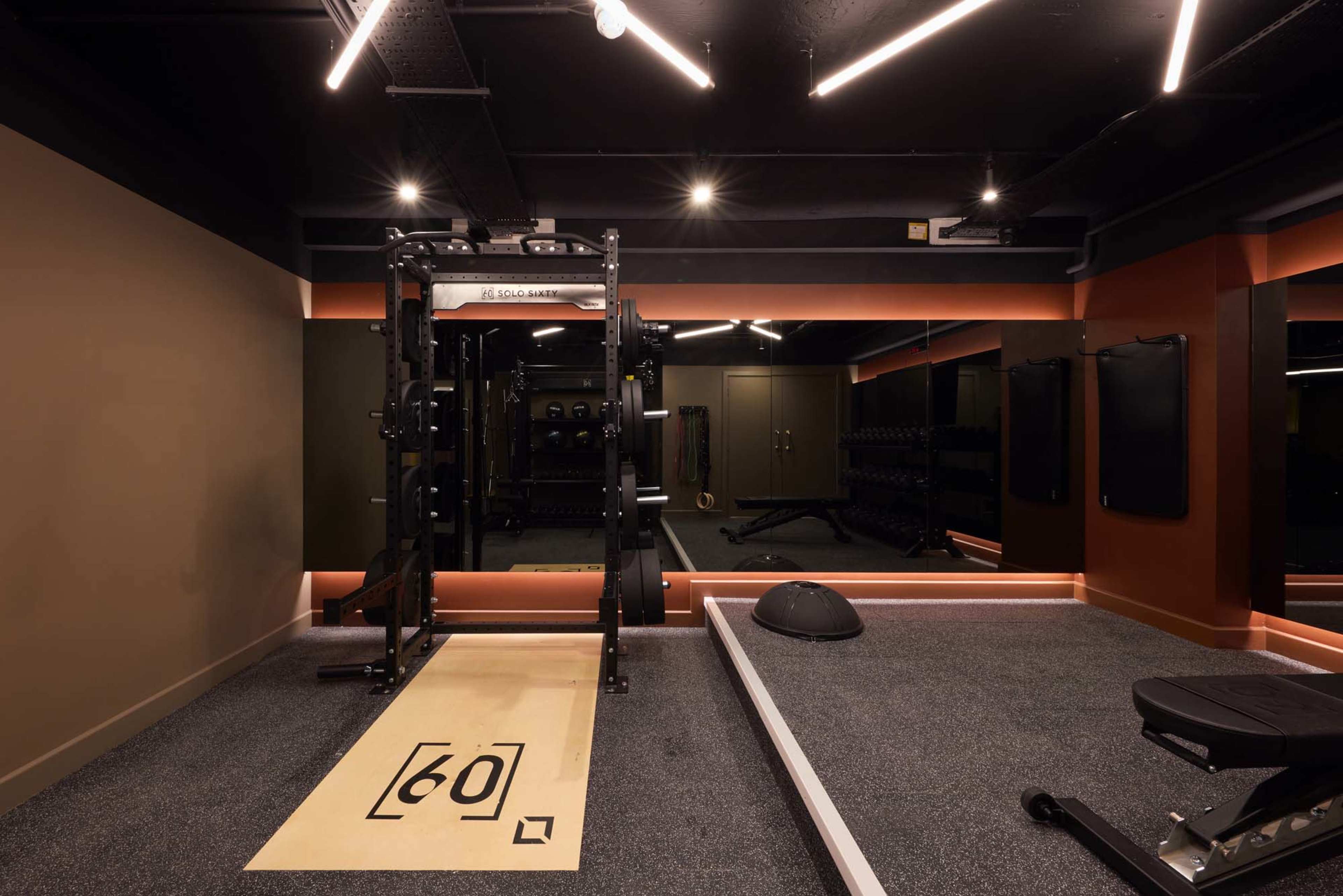 The image shows a modern gym interior equipped with weightlifting racks, free weights, and exercise benches, featuring dark walls and bright overhead lighting.