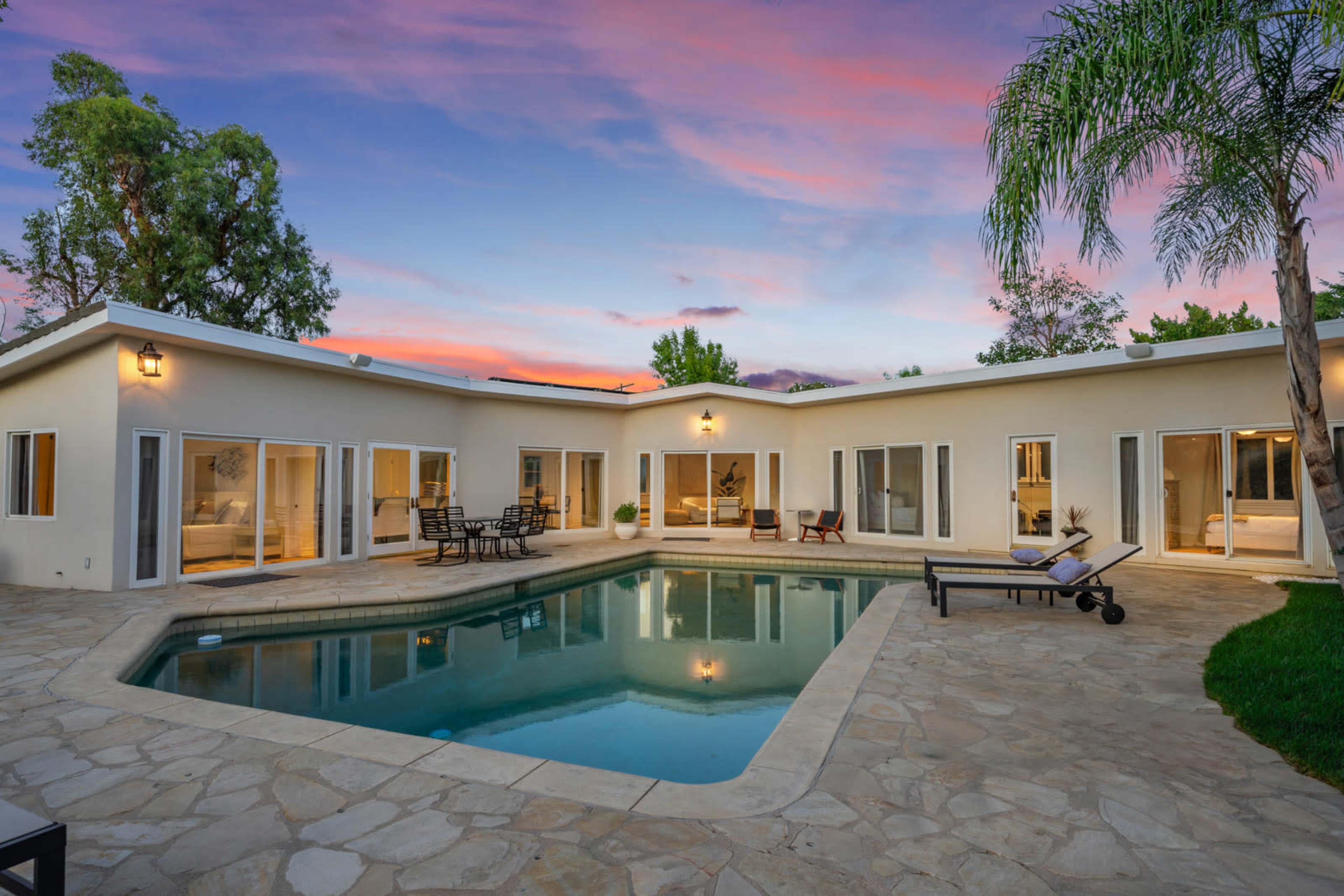 A modern single-story home features a central pool surrounded by a stone patio and large glass doors that open into the living spaces.