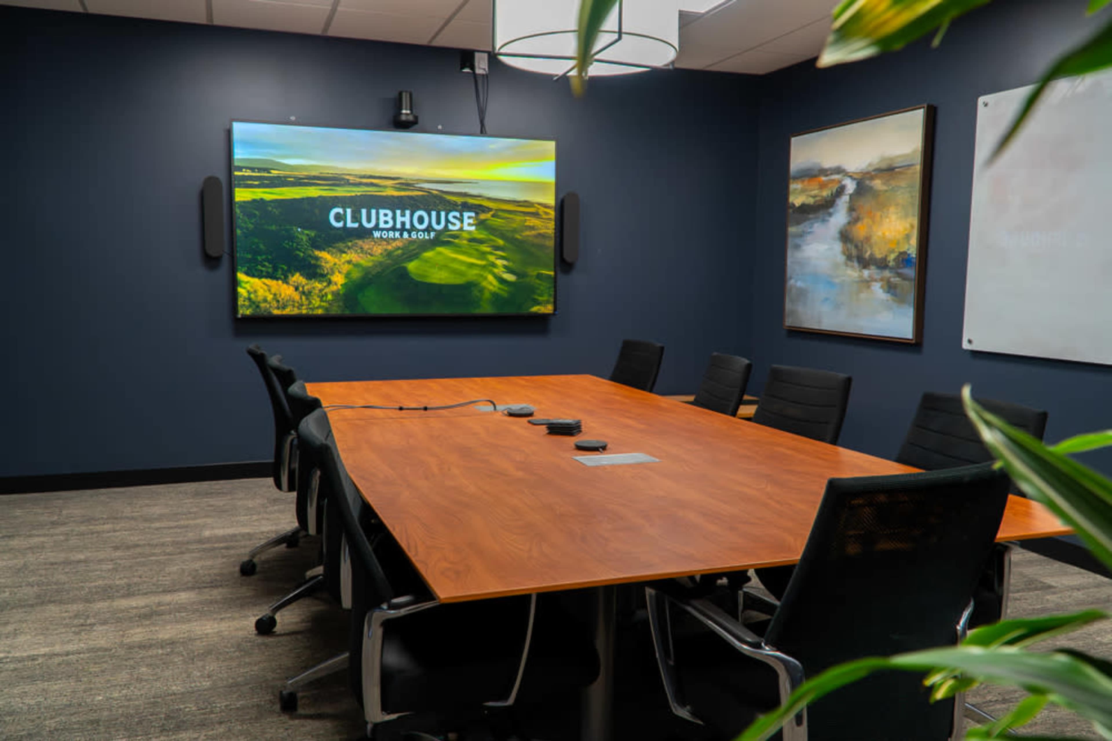 The image shows a modern conference room featuring a large wooden table, black chairs, a wall-mounted screen displaying "CLUBHOUSE NORA GOLF," and artwork on the walls.