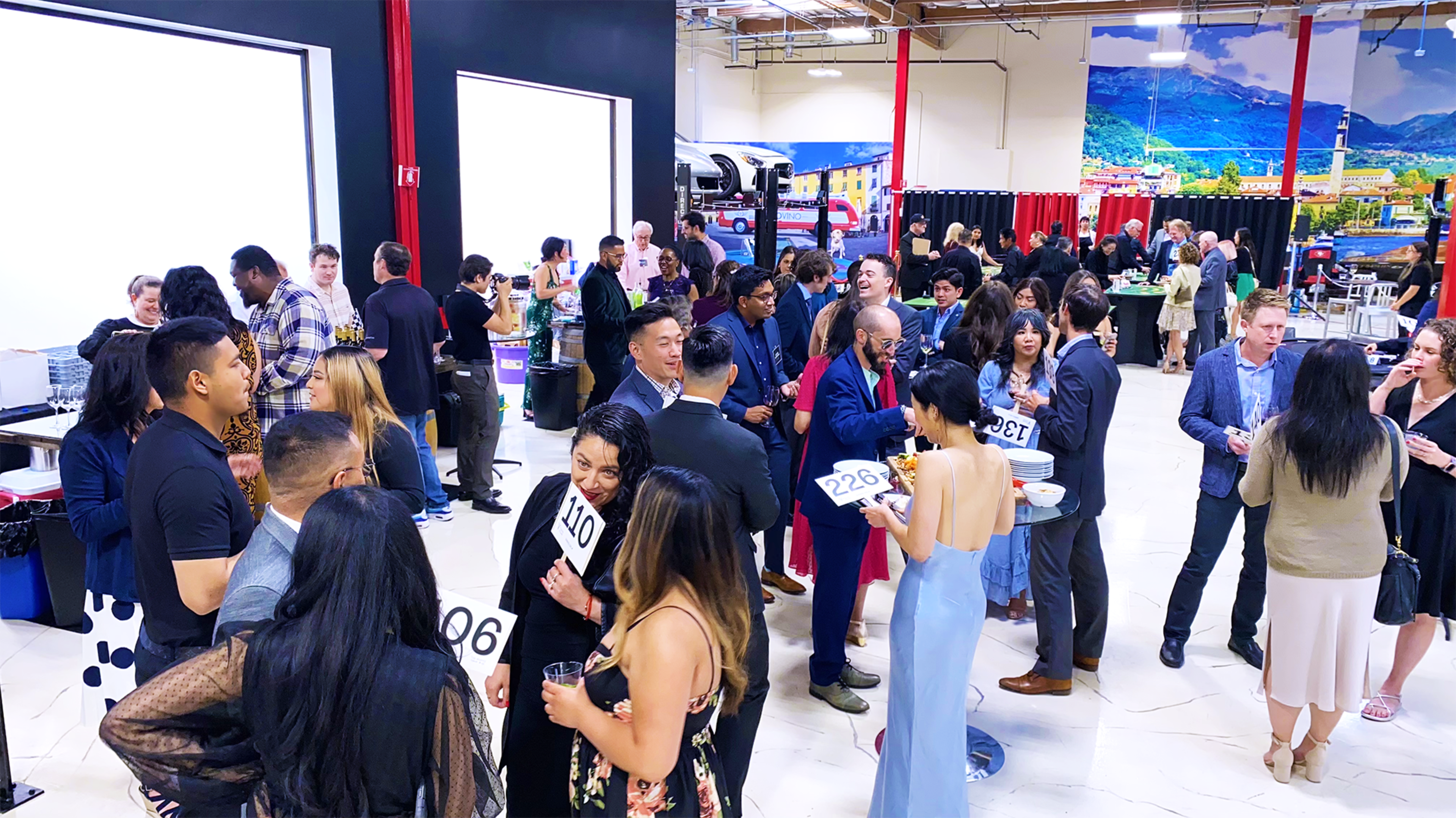 A crowded event space features attendees mingling and engaging in conversation, with some holding numbered signs and drinks.