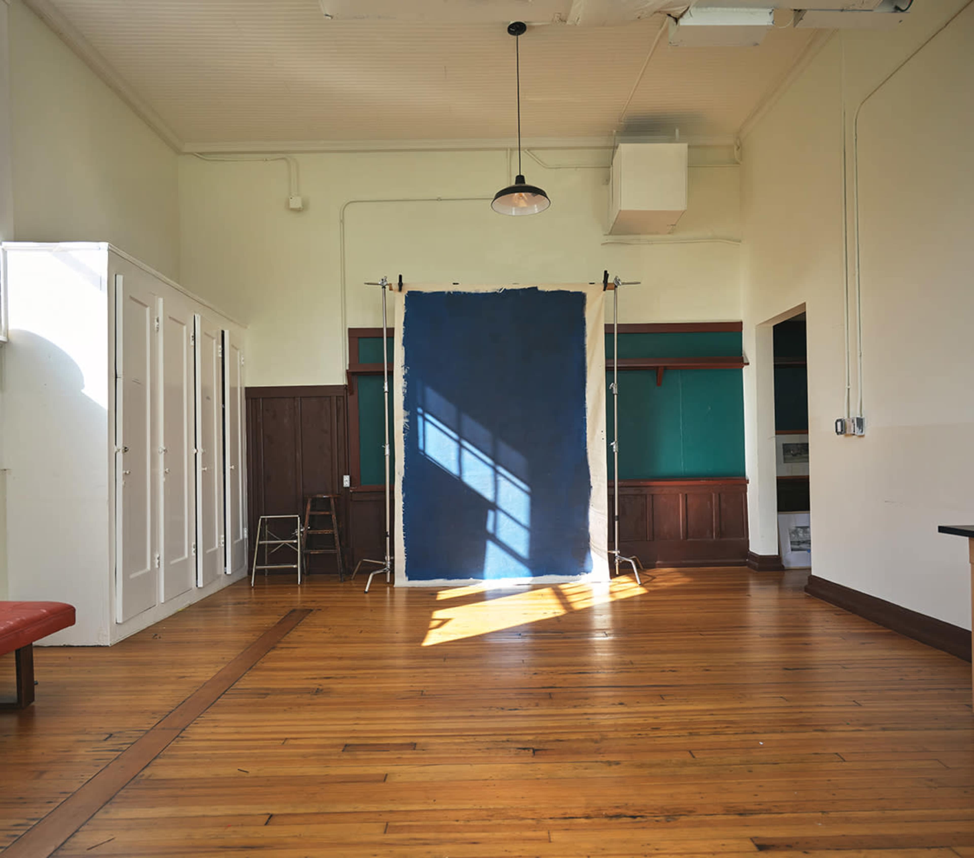 The image shows an empty room with wooden floors, a blue backdrop on stands, and sunlight streaming in through a window.