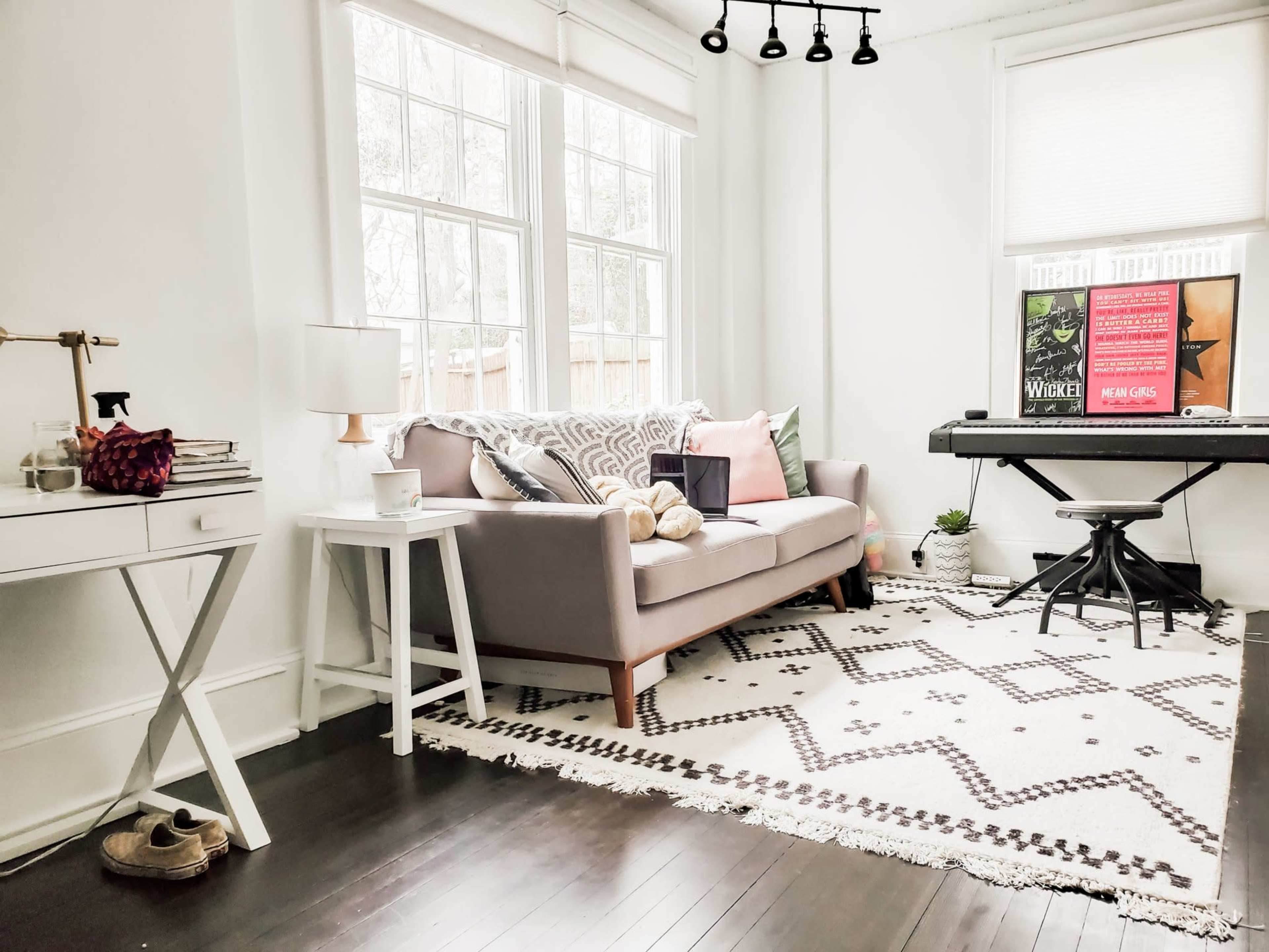 A bright, airy room featuring a light gray sofa with decorative pillows, a small table with a lamp, and a keyboard positioned next to a large window.