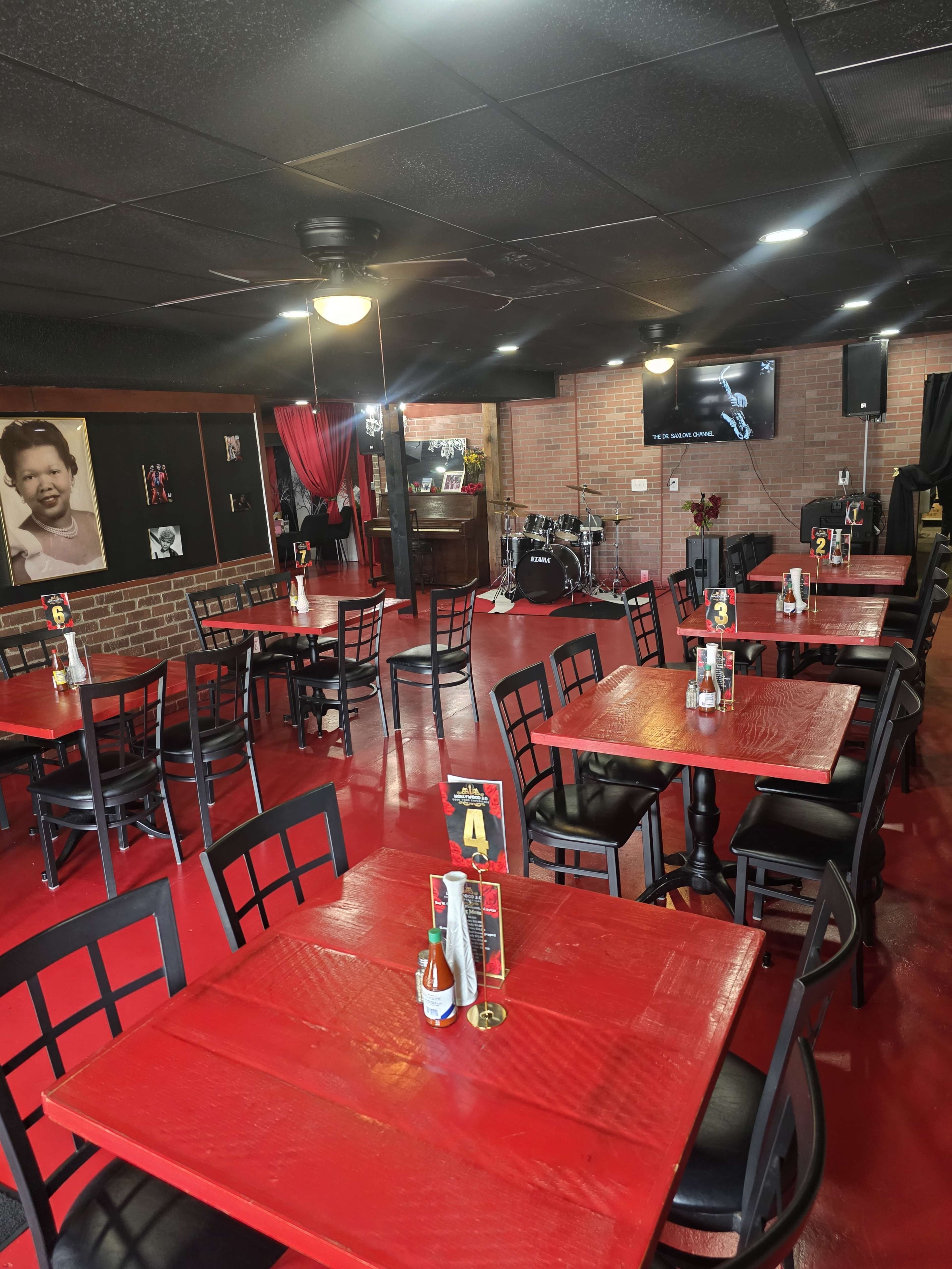The image shows a restaurant interior featuring red tables and black chairs, with a stage and drum set in the background.