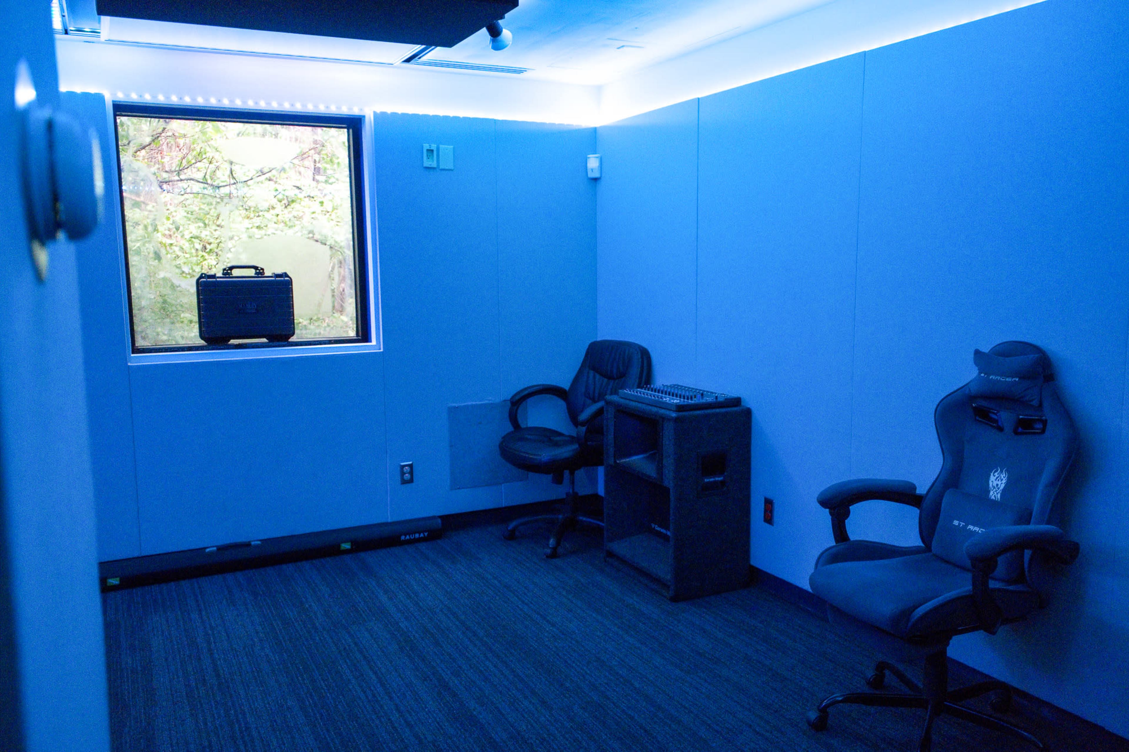 The image shows a small blue room with two chairs, a window overlooking trees, and a desk with equipment on it.