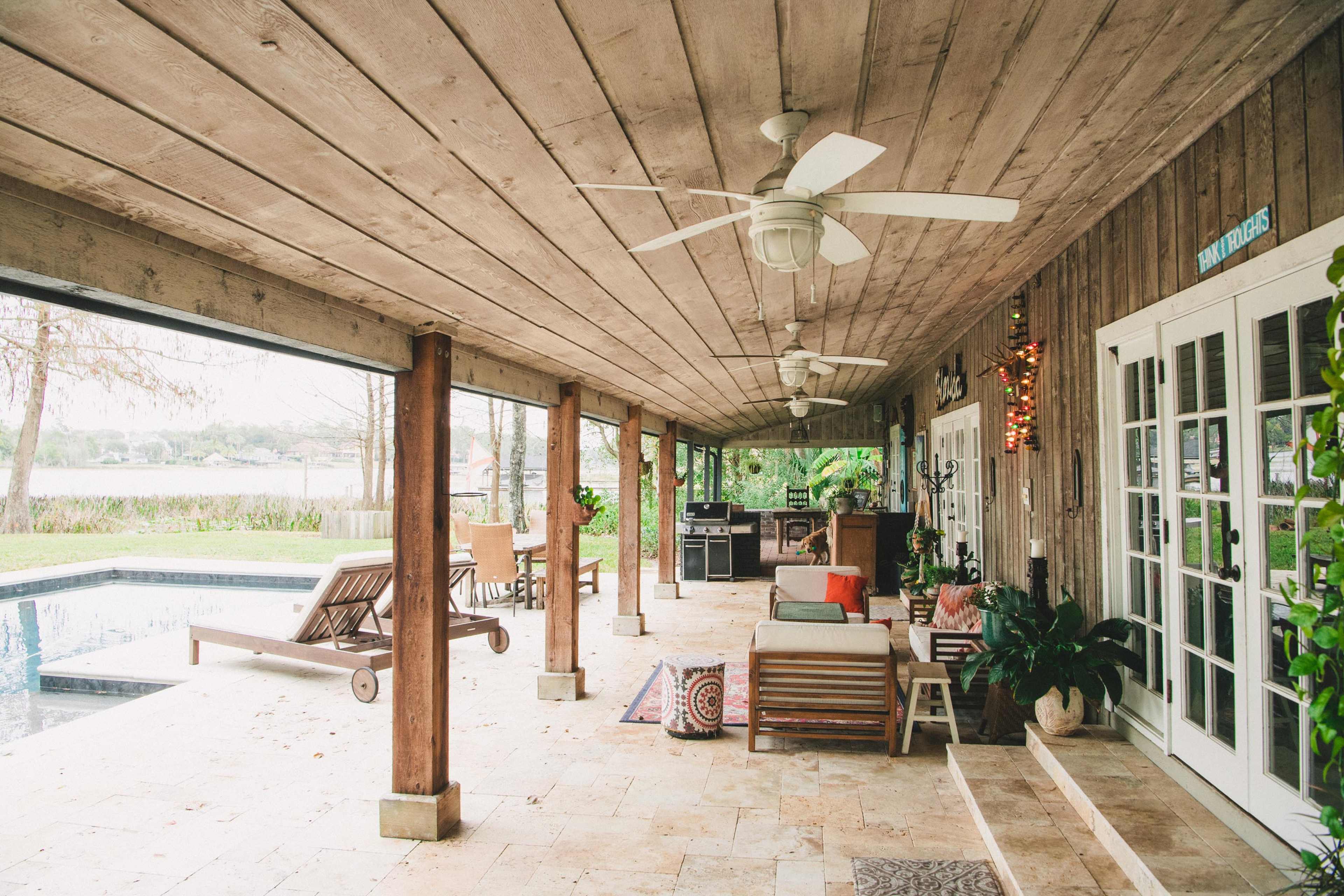 A spacious patio with wooden beams features seating, ceiling fans, and a view of a pool and lake.