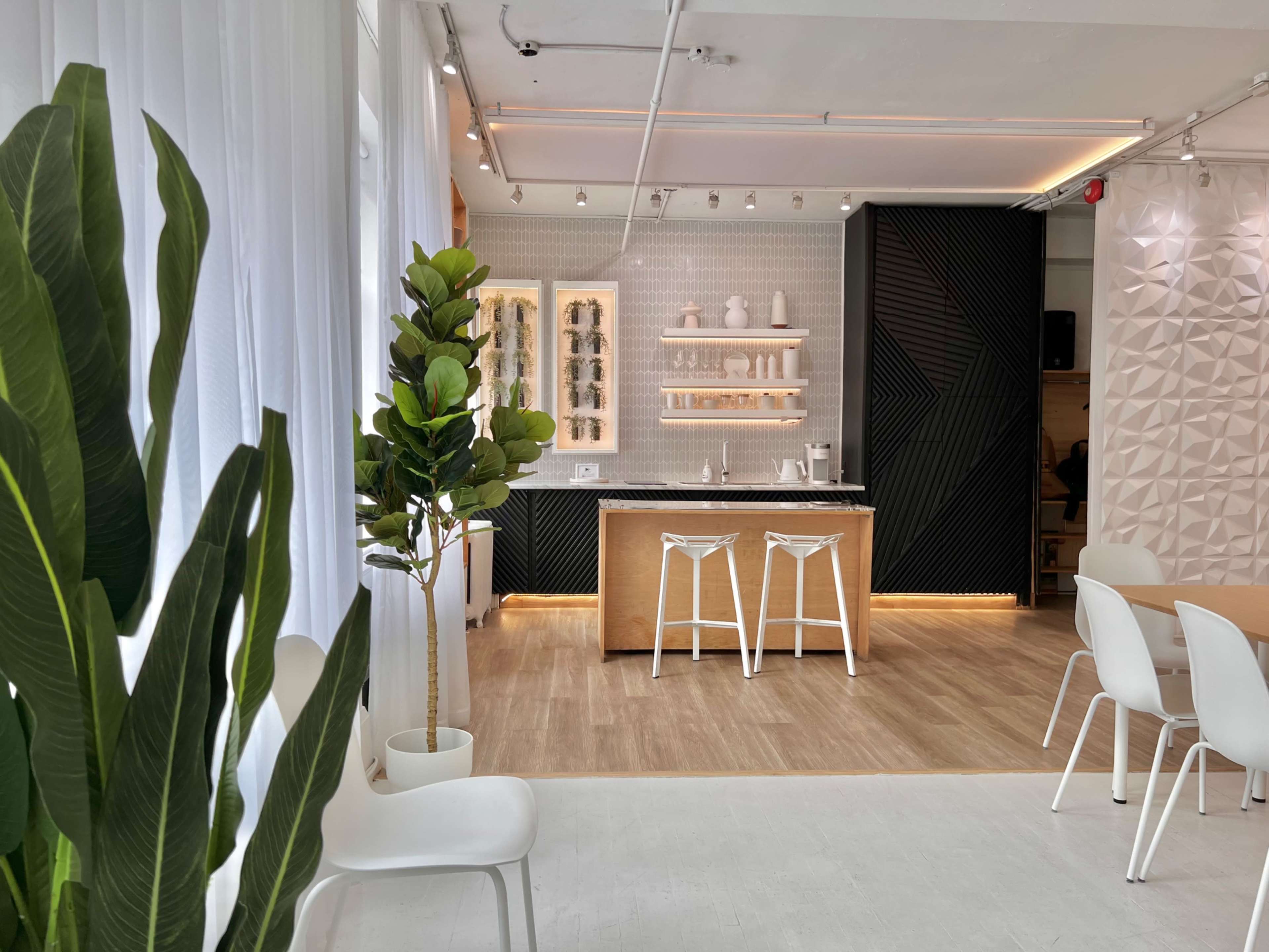 A modern interior space features a kitchen bar with stools, shelving displaying items, and greenery in a planter.
