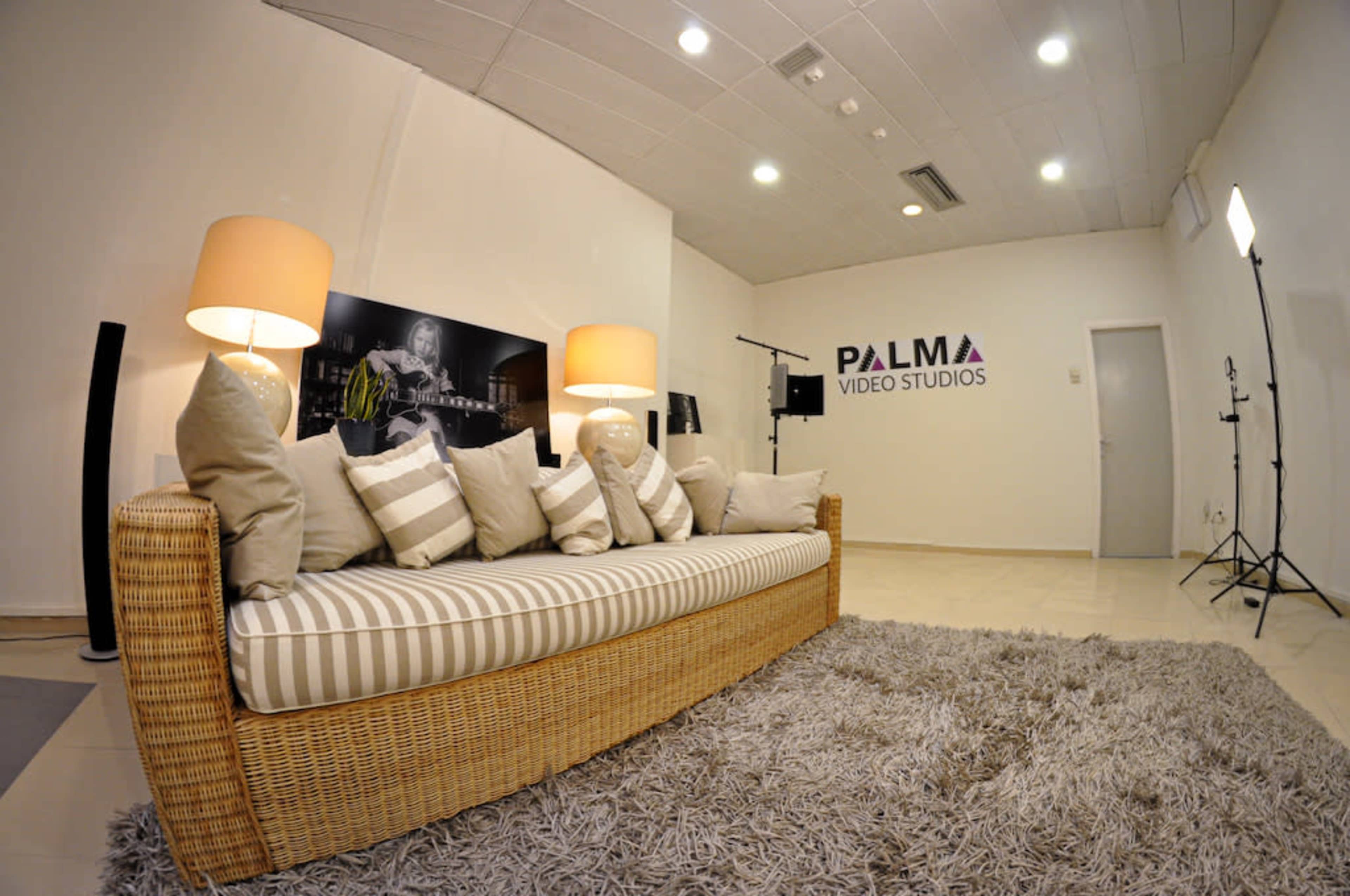 The image shows a modern video studio with a wicker sofa adorned with pillows, a plush gray rug, and studio lighting equipment.