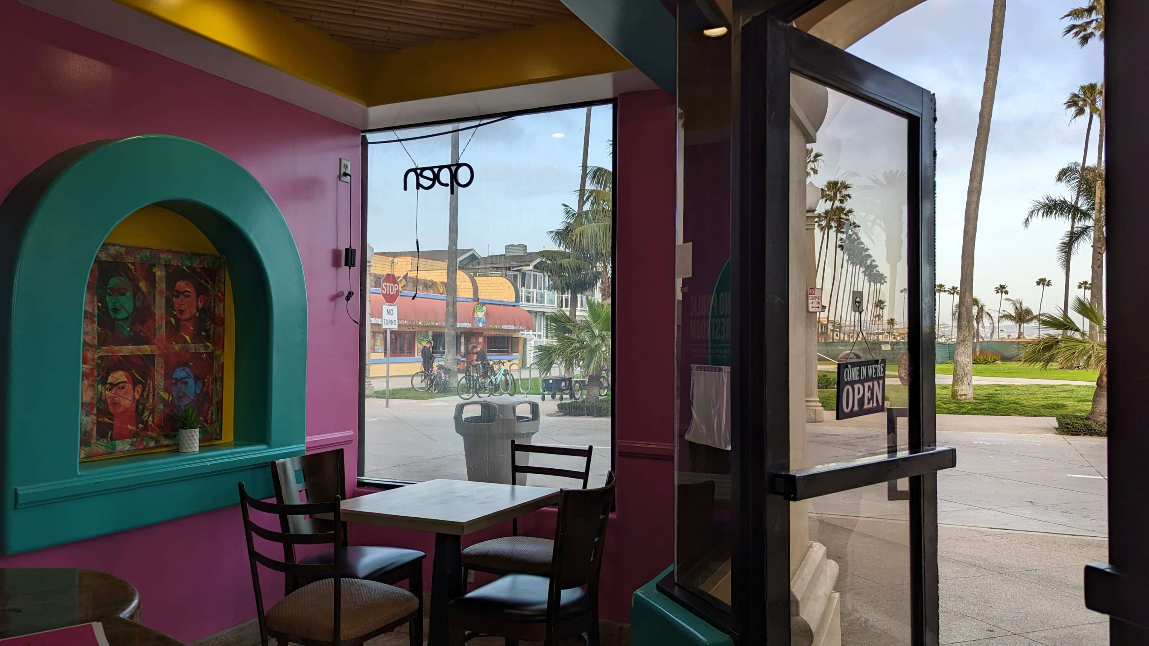 A colorful cafe interior with a view of a palm-lined street and shops through large glass doors.
