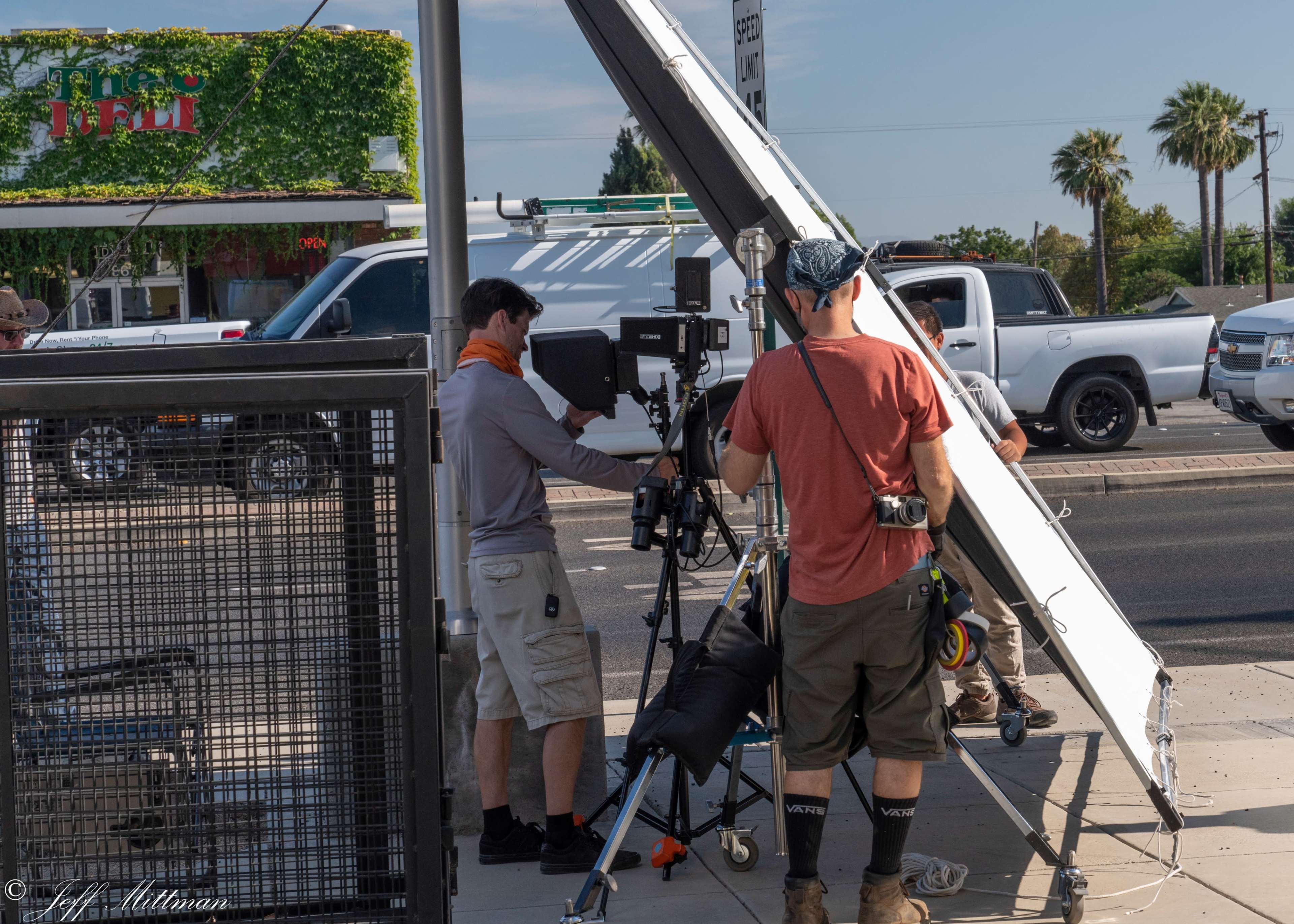 A film crew sets up equipment on a sidewalk near a restaurant and palm trees under clear skies.