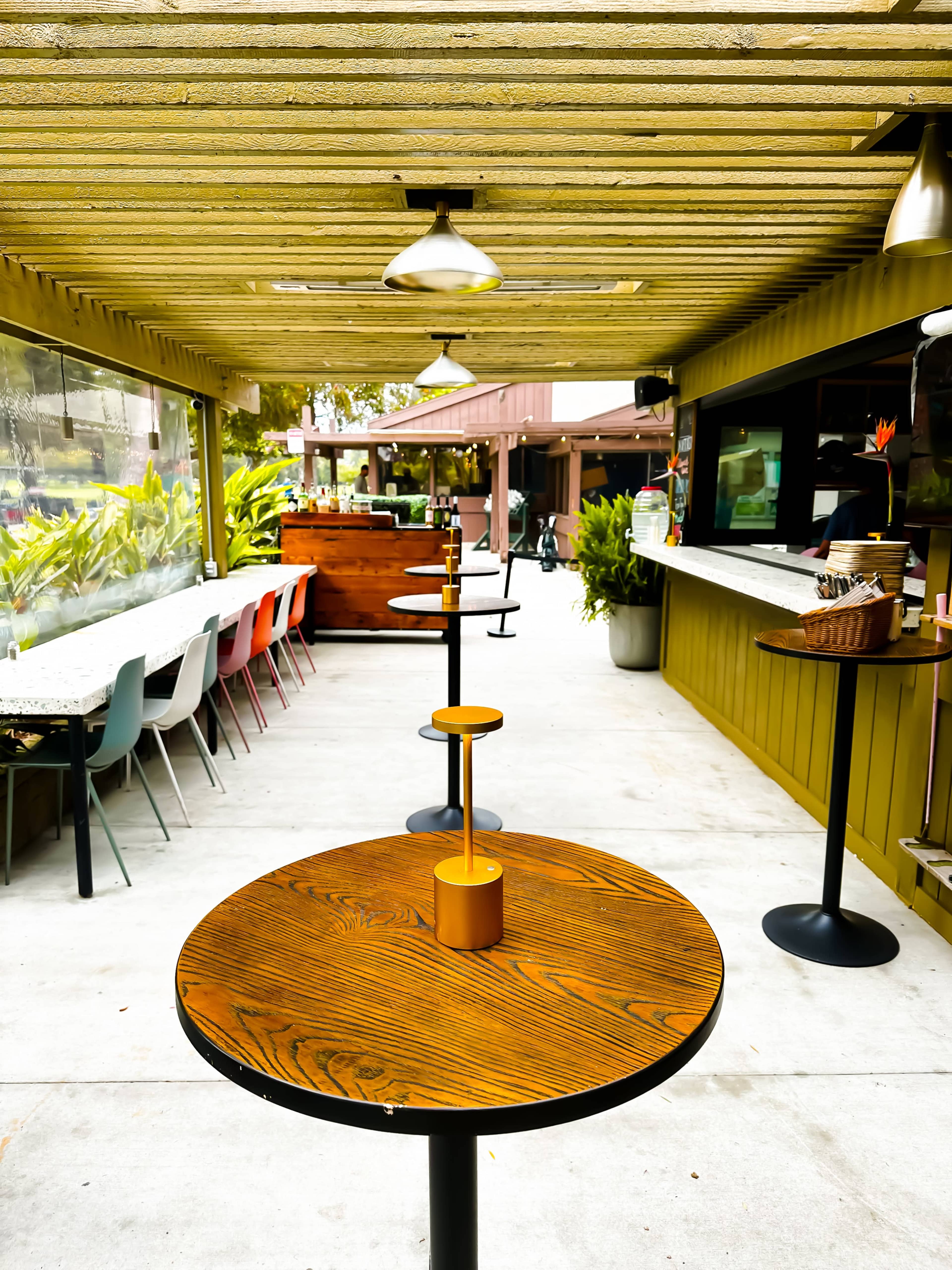 The image shows a modern outdoor café space with wooden tables, a counter, and colorful chairs lined along the sides.
