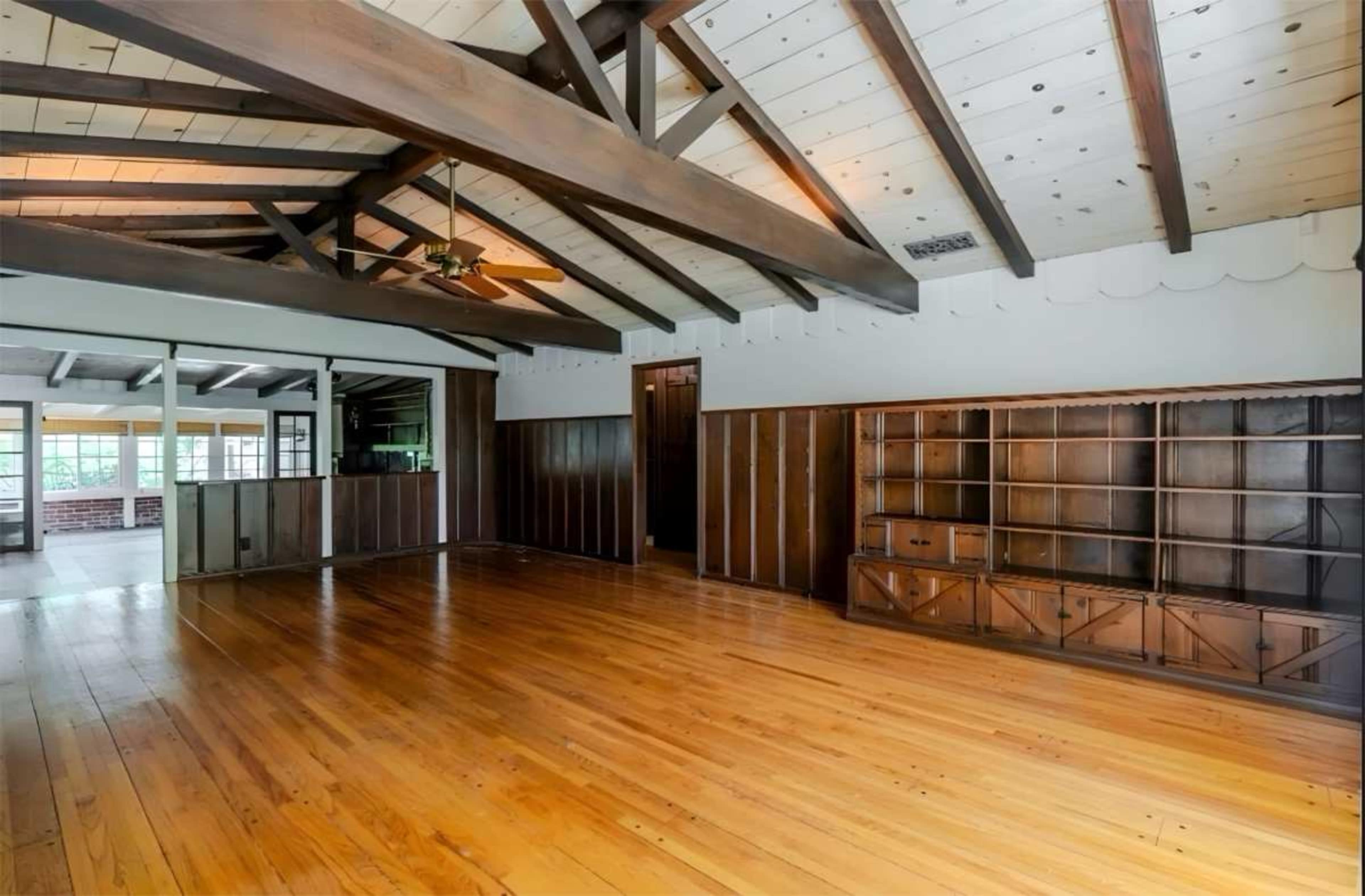 A large, empty living room features exposed wooden beams, a ceiling fan, and hardwood floors, with built-in shelving against one wall.