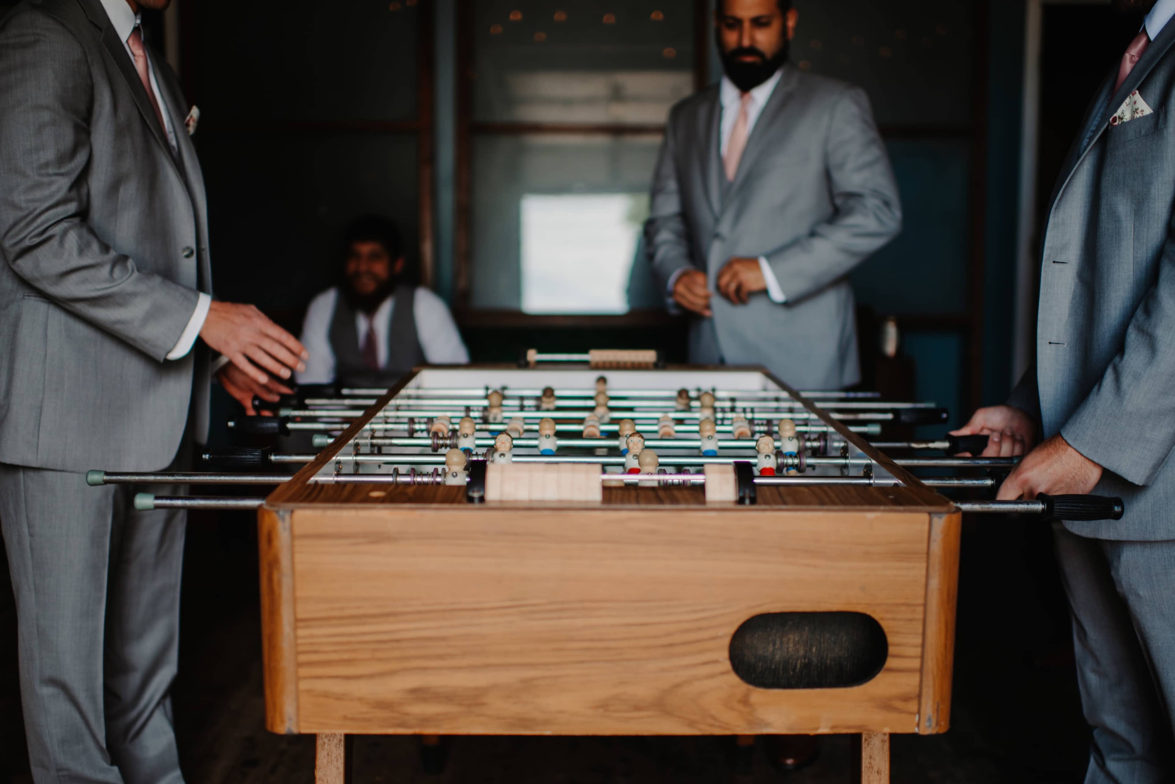 Two men in suits play foosball while a third man observes in the background.