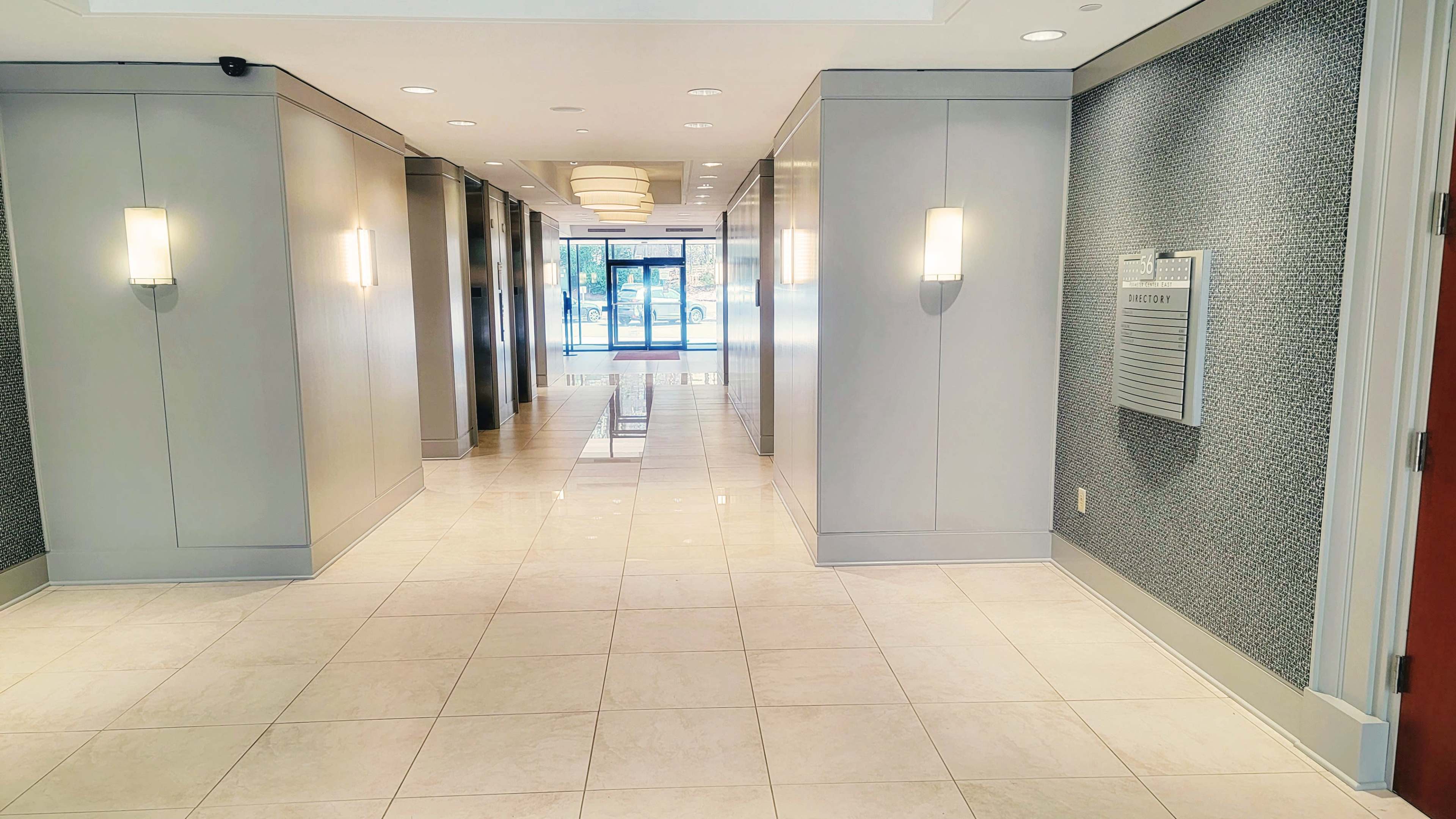 A modern corridor with glossy tile flooring, flanked by grey wall panels and illuminated by wall sconces, leading to a glass entrance.