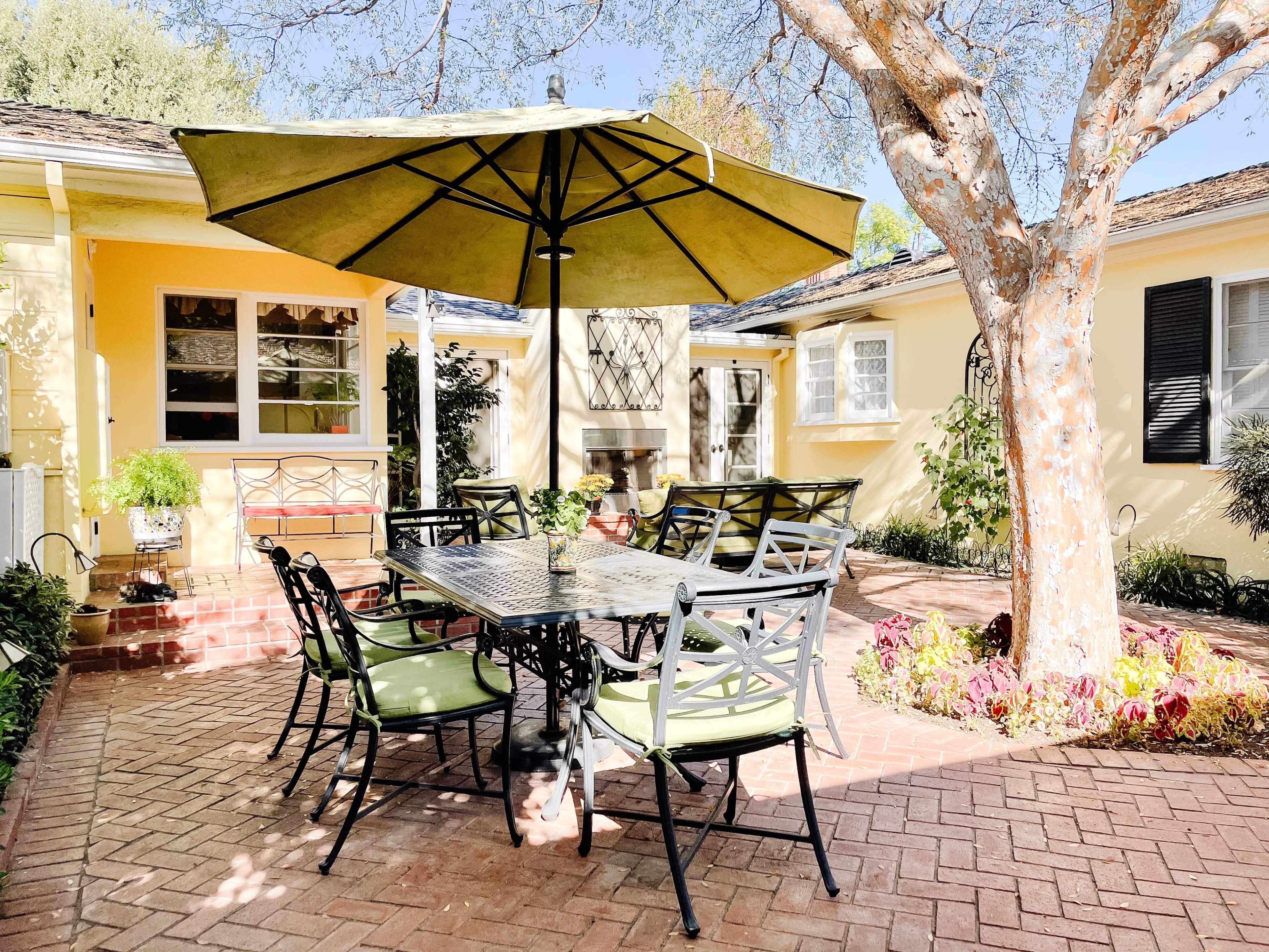 A patio area features a table and chairs under a large umbrella, surrounded by greenery and a cozy yellow house.