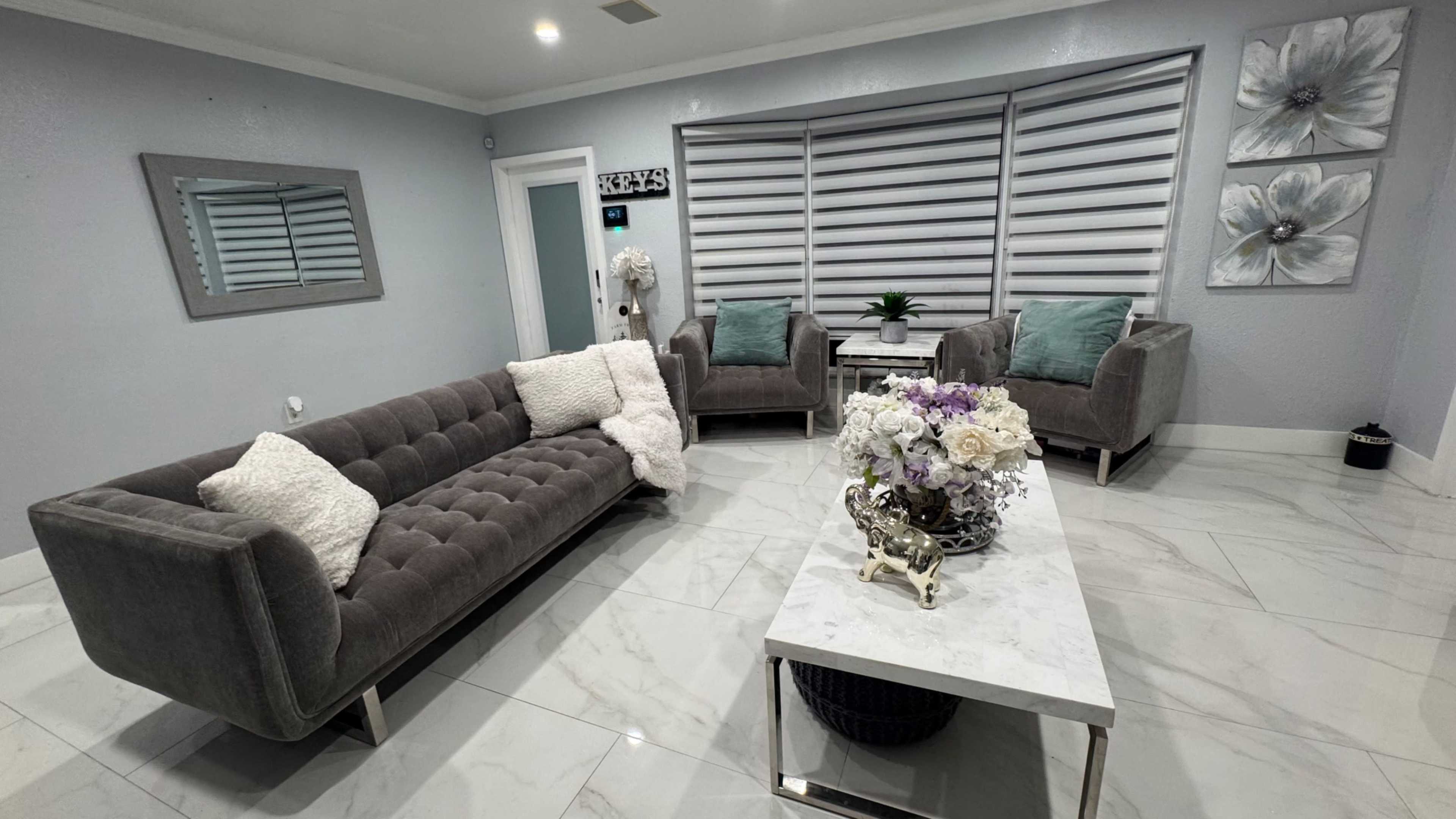 The image shows a modern living room with a gray tufted sofa, two armchairs with cushions, a marble coffee table, and floral decorations.
