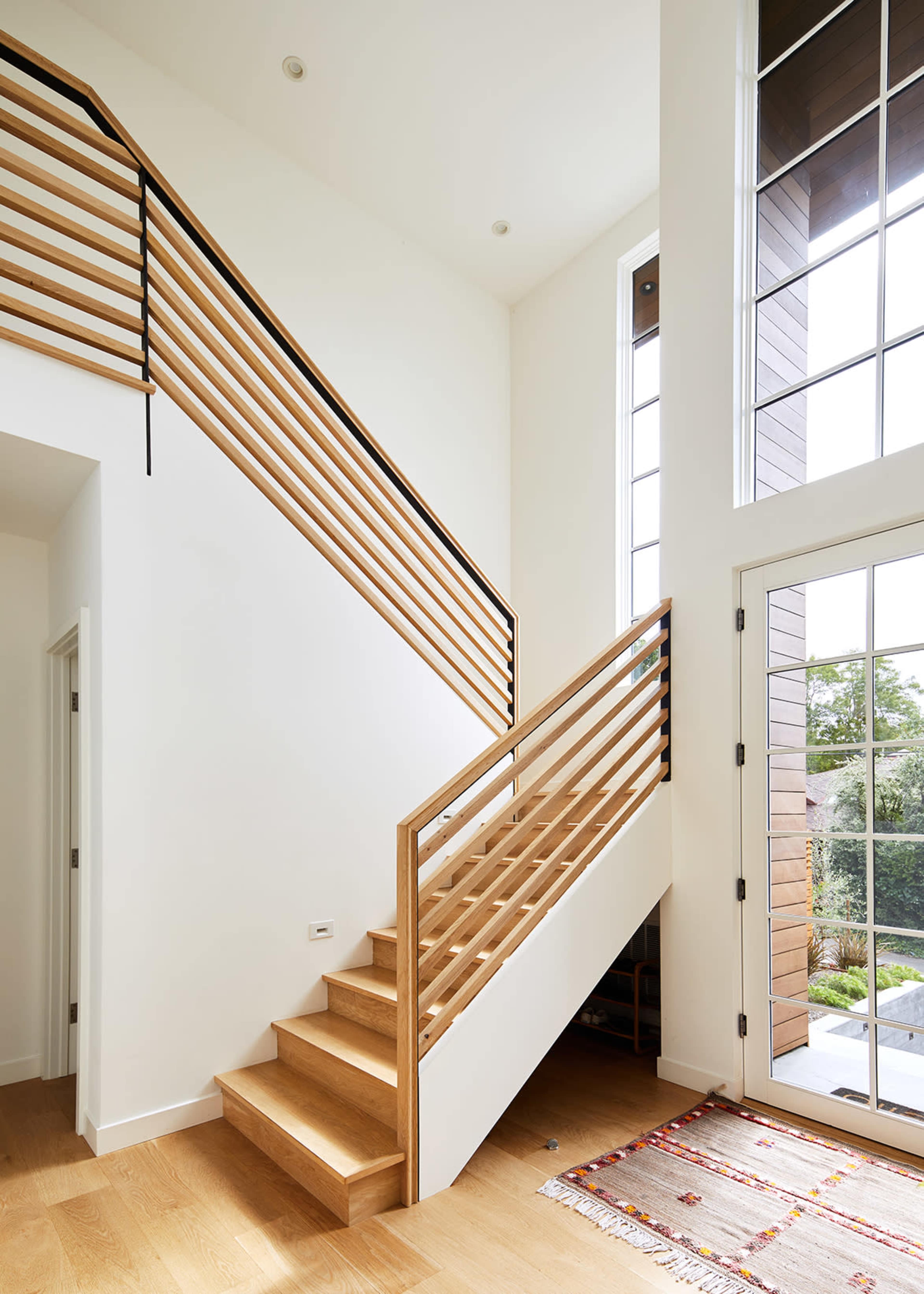 The image shows a modern staircase with wooden steps and a wooden railing, leading to a second floor in a bright entryway with large windows.