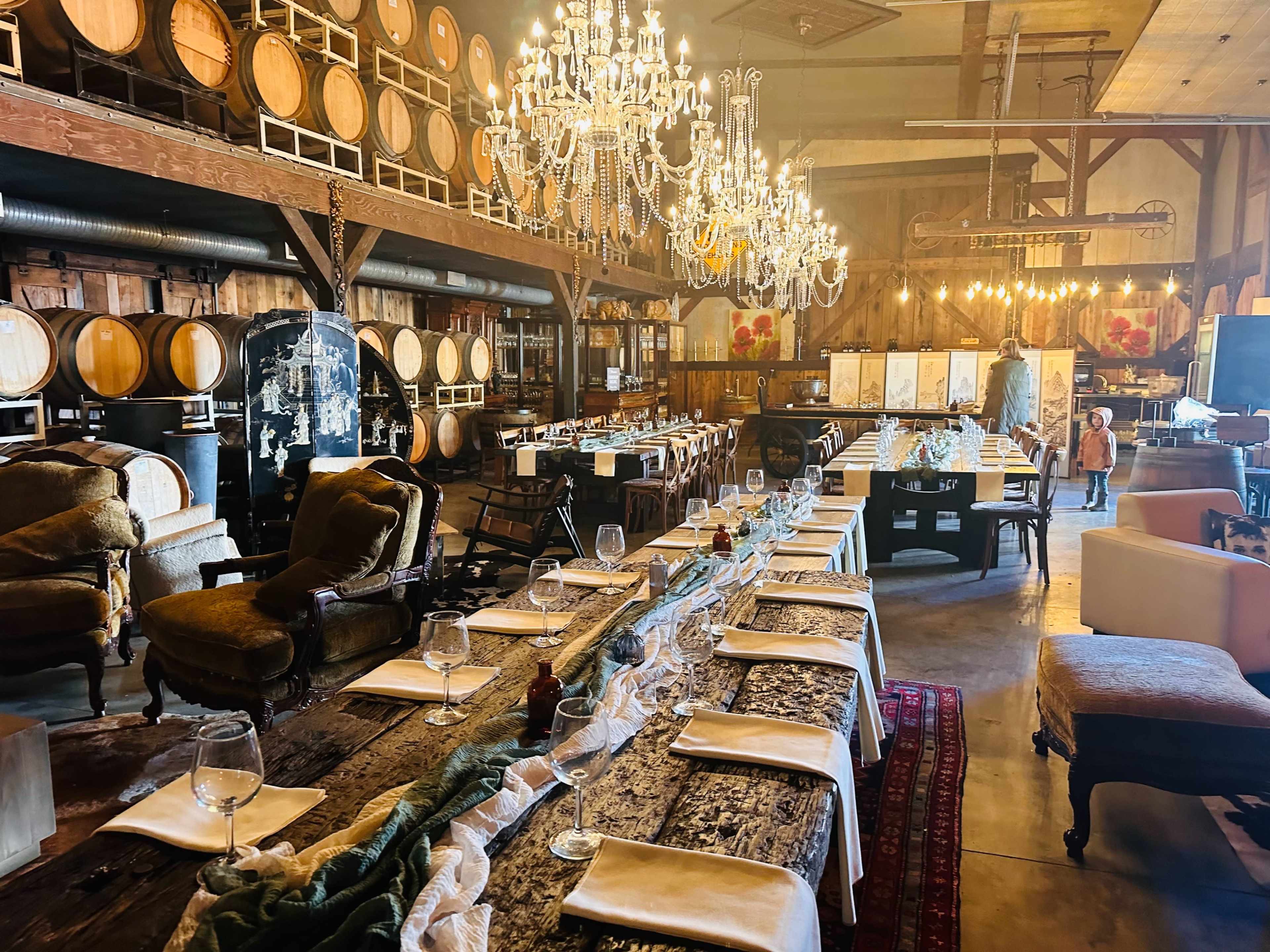 A large dining room is set with a long wooden table surrounded by chairs, illuminated by chandeliers, with wine barrels and rustic decor in the background.