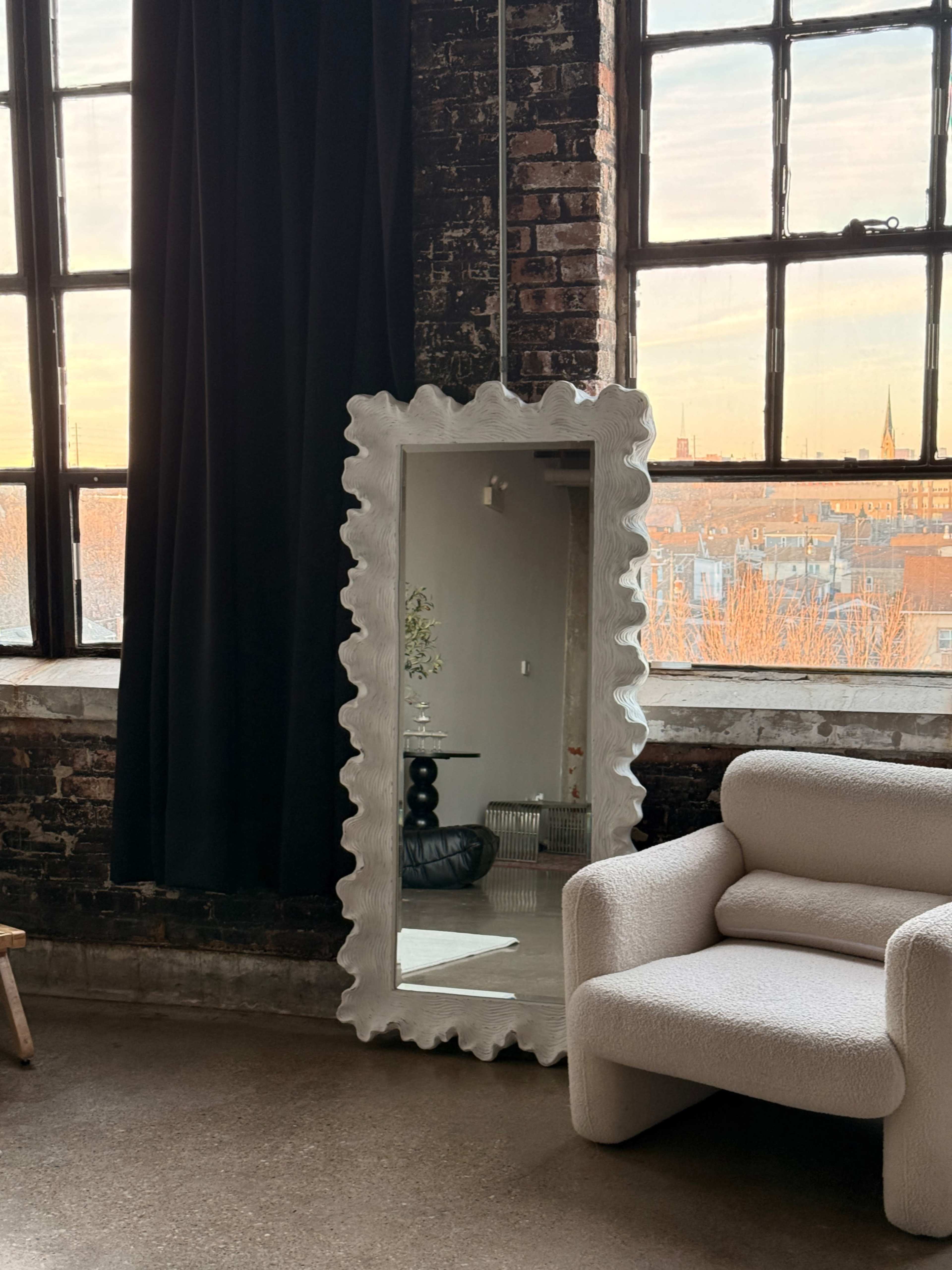 A large, irregularly shaped mirror stands against a brick wall near a window, reflecting the light from the sunset, with a cozy, white armchair nearby.