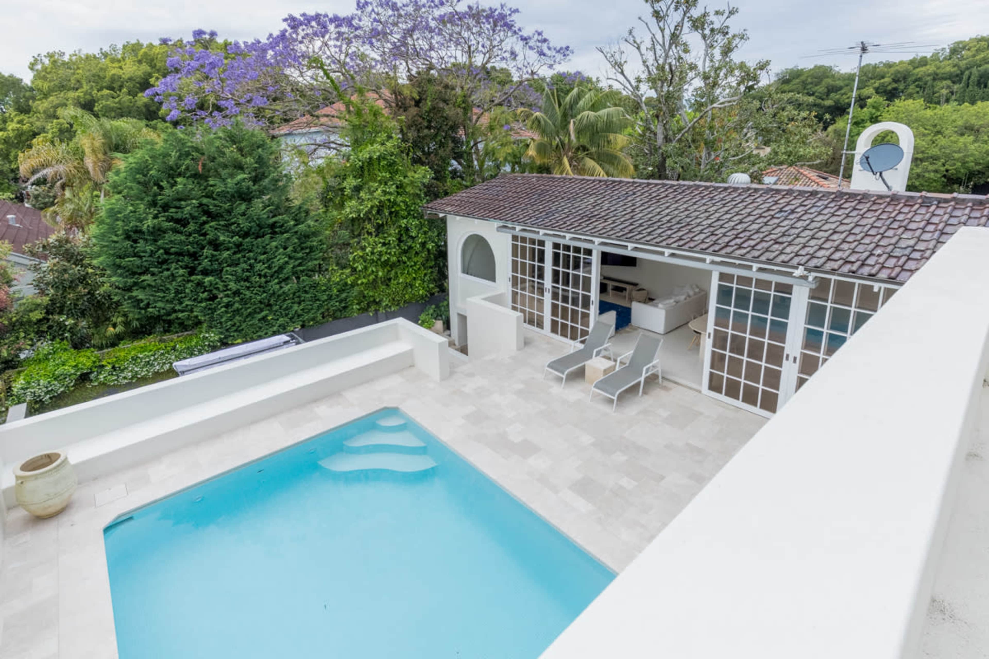 A modern pool surrounded by a stone deck, with a white building featuring large windows and a roof overhang in the background.