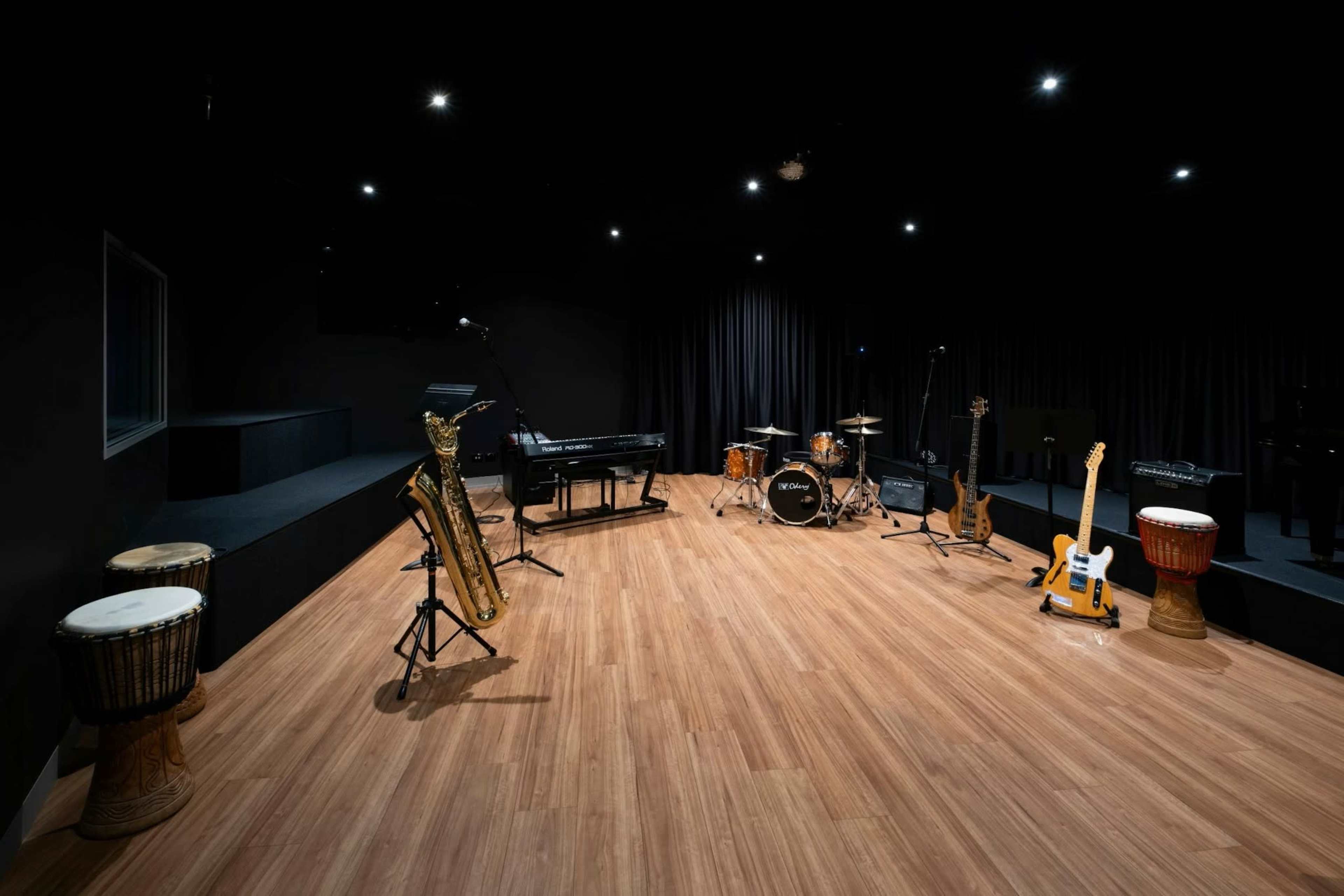 The image shows a music studio with various instruments, including a saxophone, drums, a keyboard, guitars, and percussion instruments, arranged on a polished wooden floor under dim lighting.